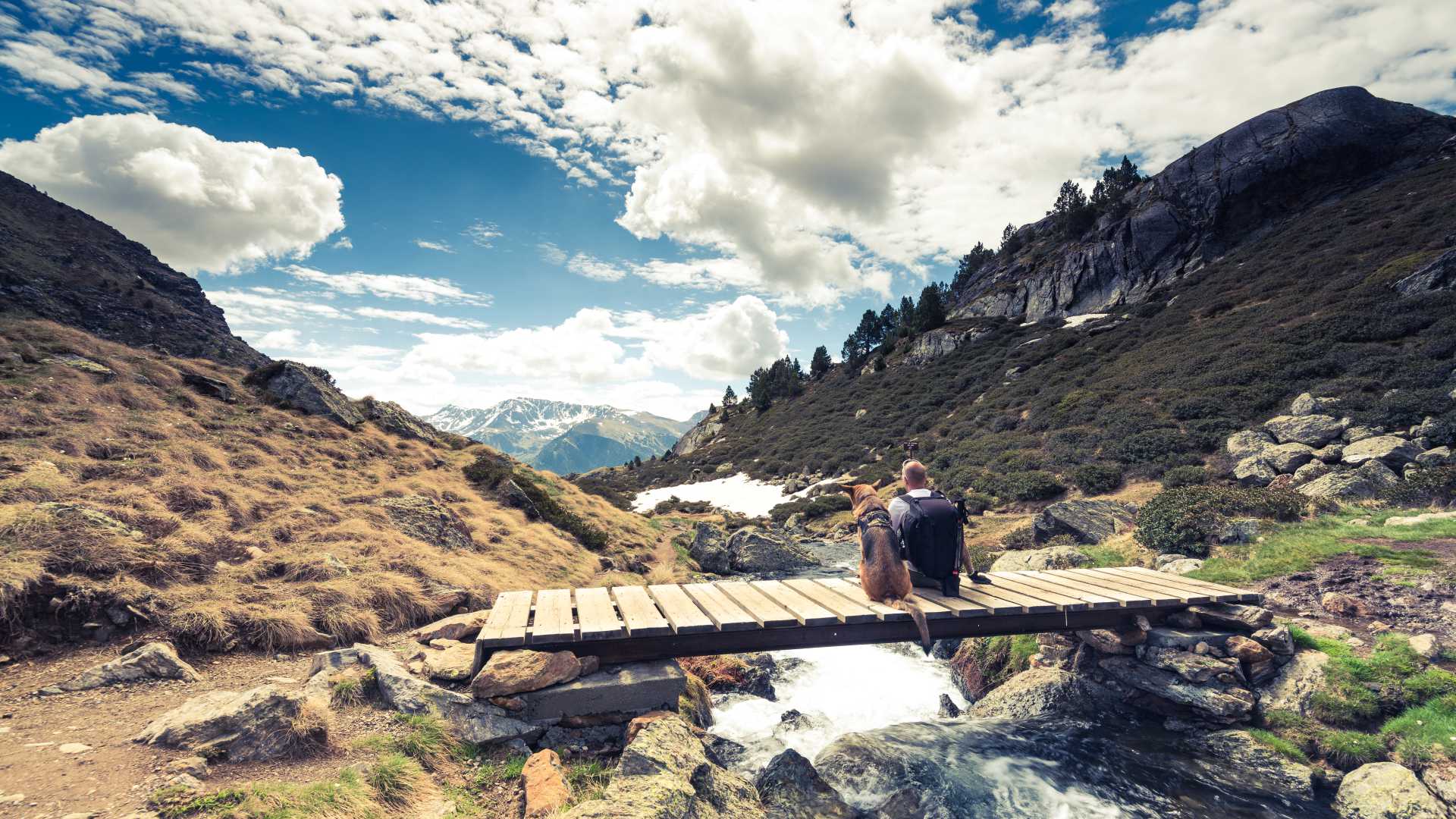 Ein Wanderer und sein Hund ruhen sich auf einer Holzbrücke aus und genießen unter einem strahlend blauen Himmel den malerischen Bergblick in Andorra.