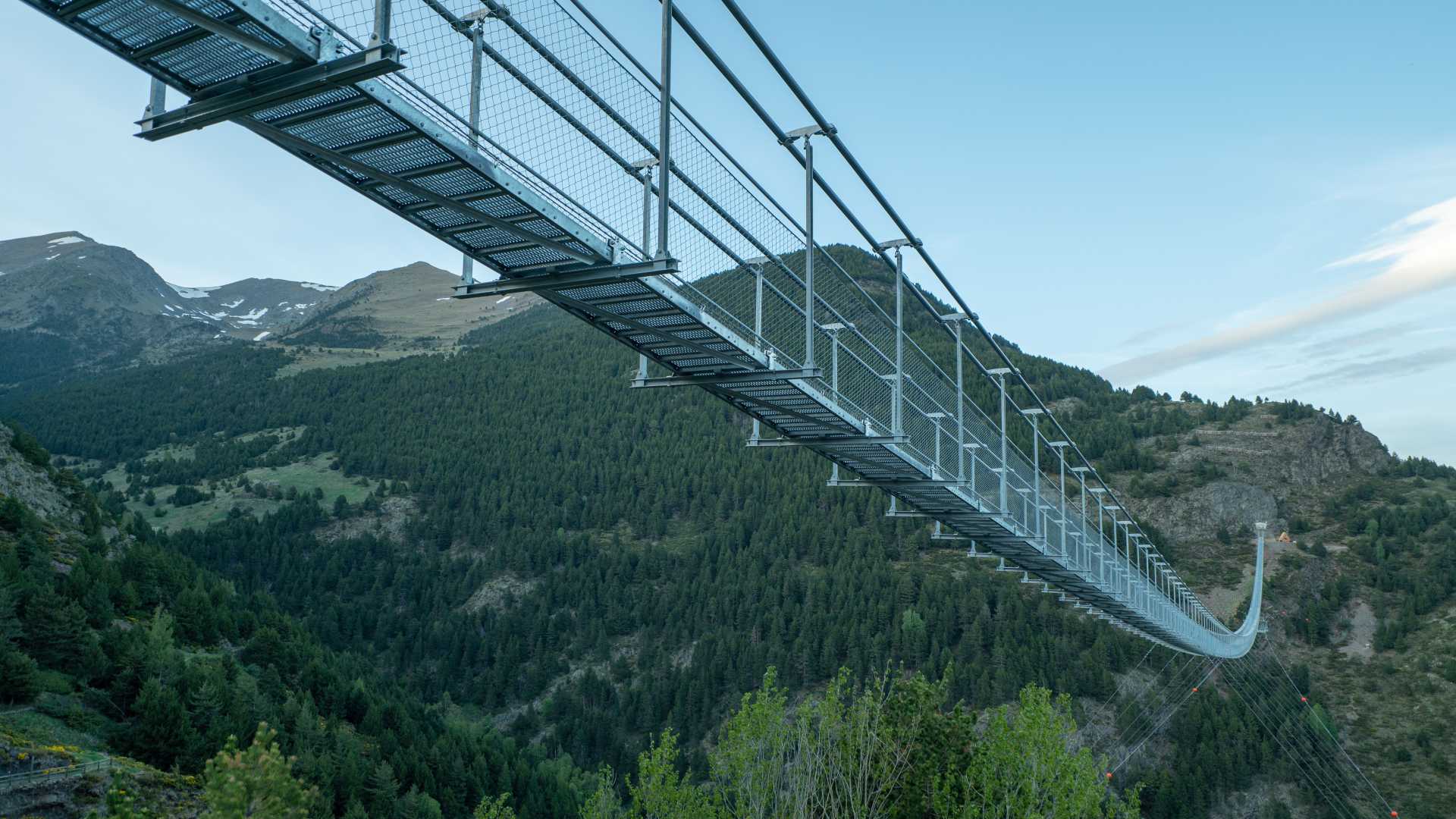 Eine tibetische Metallbrücke überspannt ein üppiges grünes Tal in Canillo, Andorra, mit bewaldeten Bergen im Hintergrund.