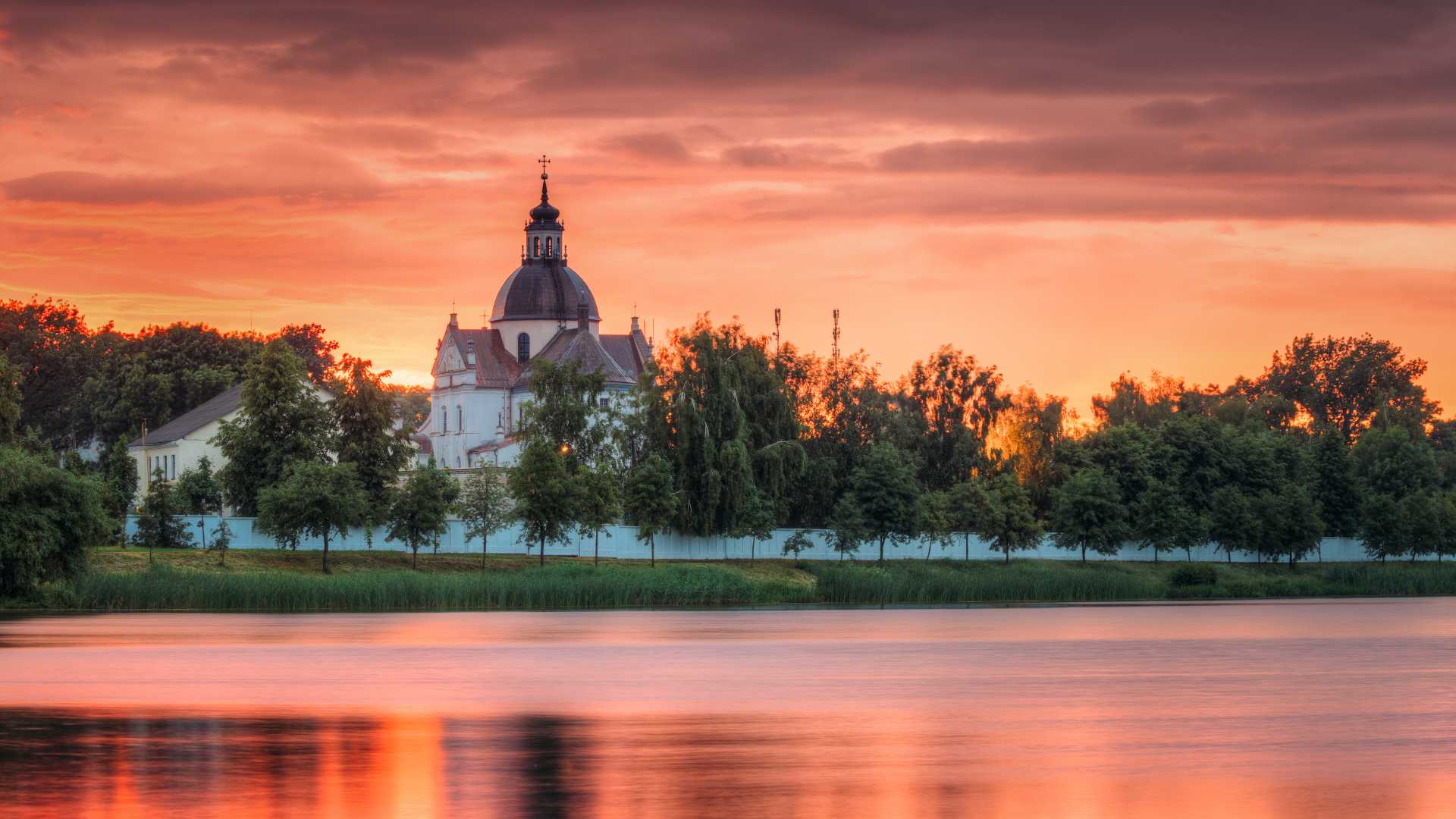 De Corpus Christi-kerk bij een stil meer bij zonsondergang in Nesvizh, Wit-Rusland, met een levendige lucht die op het water reflecteert.