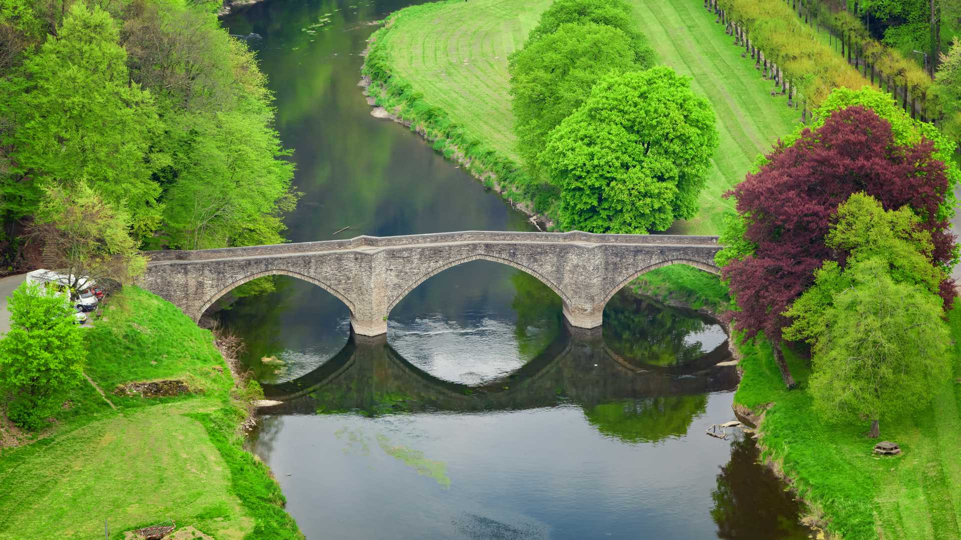 High-angle view of Pont de Cordemoy, an arch stone bridge over the Semois River in Bouillon, Belgium, surrounded by greenery.