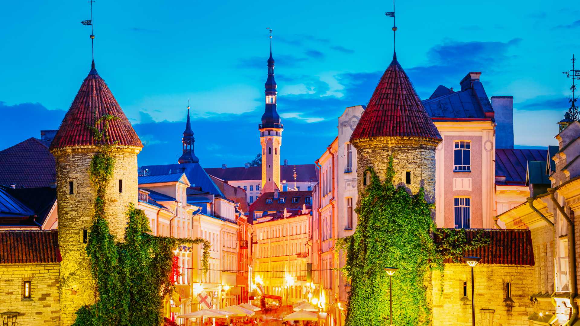 Illuminated Viru Gate in Tallinn's Old Town at night, featuring medieval towers and vibrant street life.