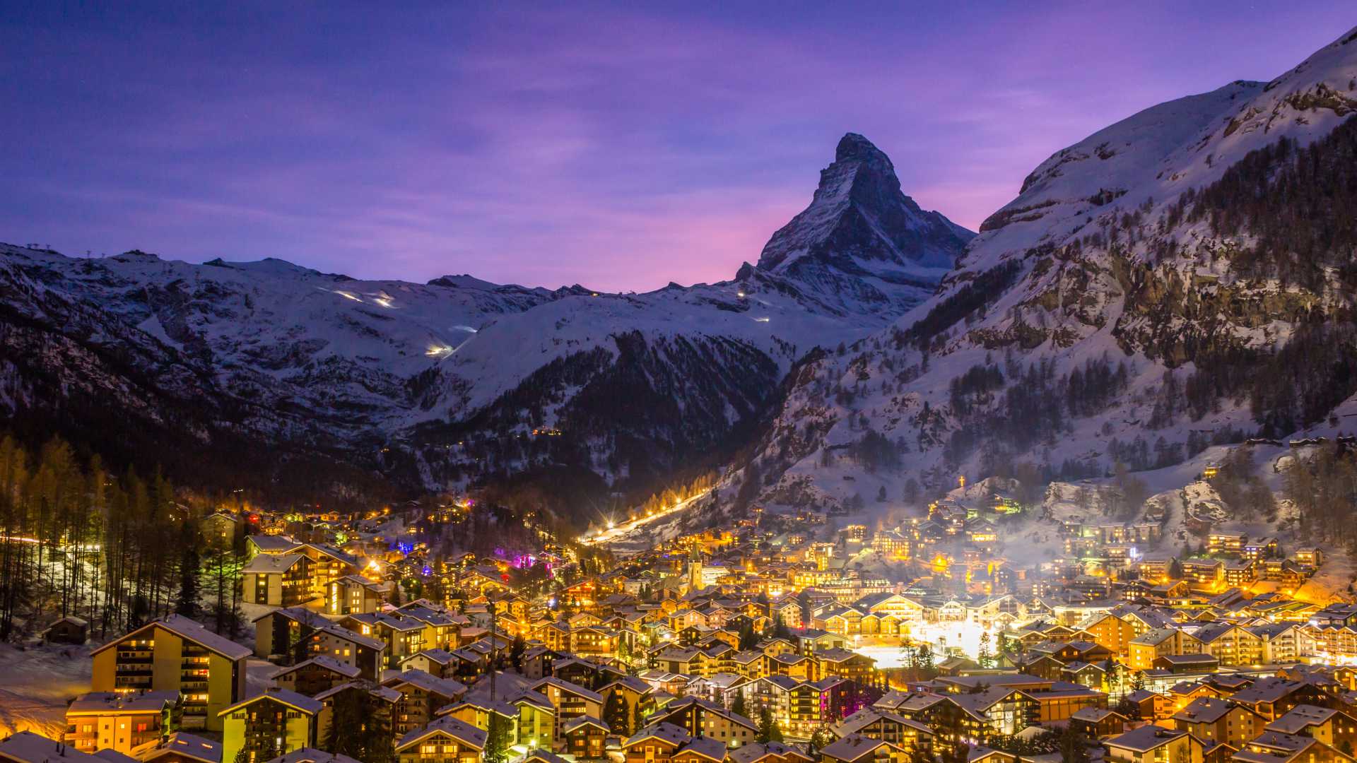 Zermatts gemütliche Lichter funkeln unter einem violetten Himmel, mit dem ikonischen Matterhorn, das sich hoch in den Schweizer Alpen erhebt.