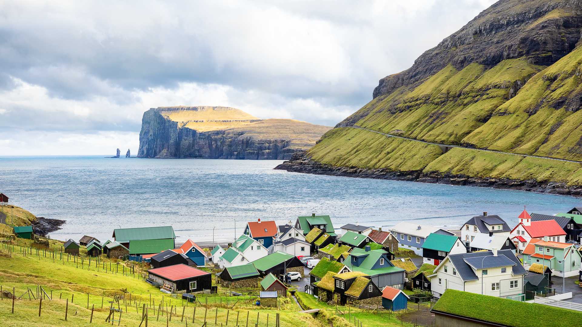 Colorful houses of Tjørnuvík village beneath dramatic cliffs, with the sea stacks Risin and Kellingin in the distance, Faroe Islands.
