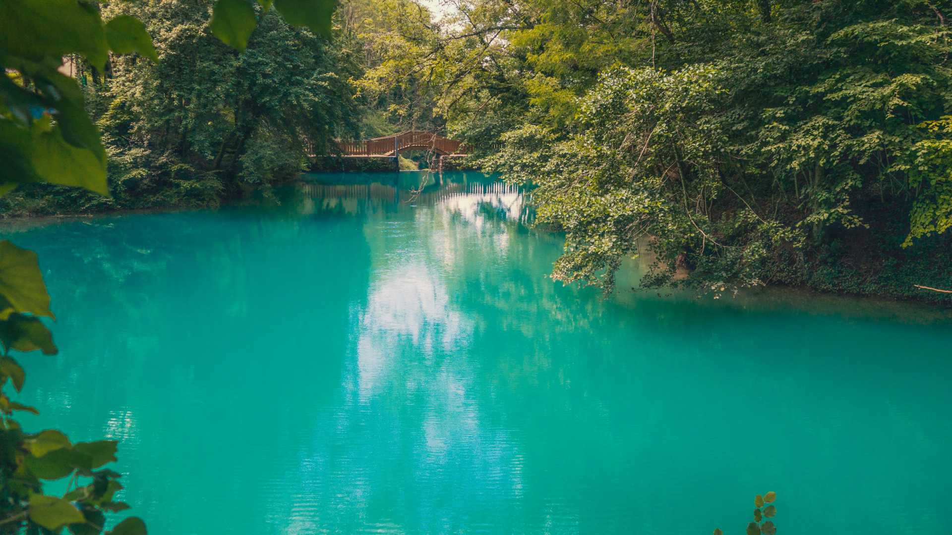 Een levendig turquoise meer omgeven door groen, met een schilderachtig houten bruggetje.