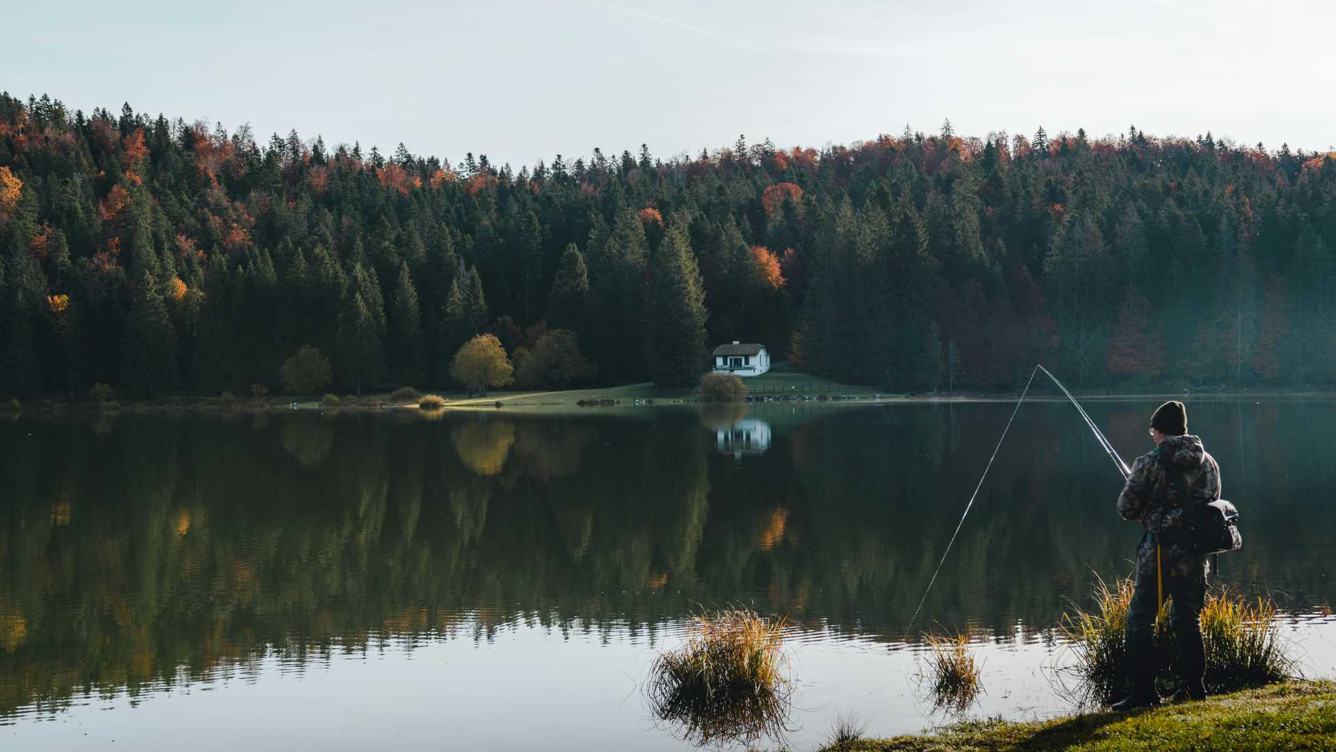 Een sereen meer met een eenzame visser omringd door herfstbomen en een klein huis aan het meer bij Lac Genin, Oyonnax, Frankrijk.