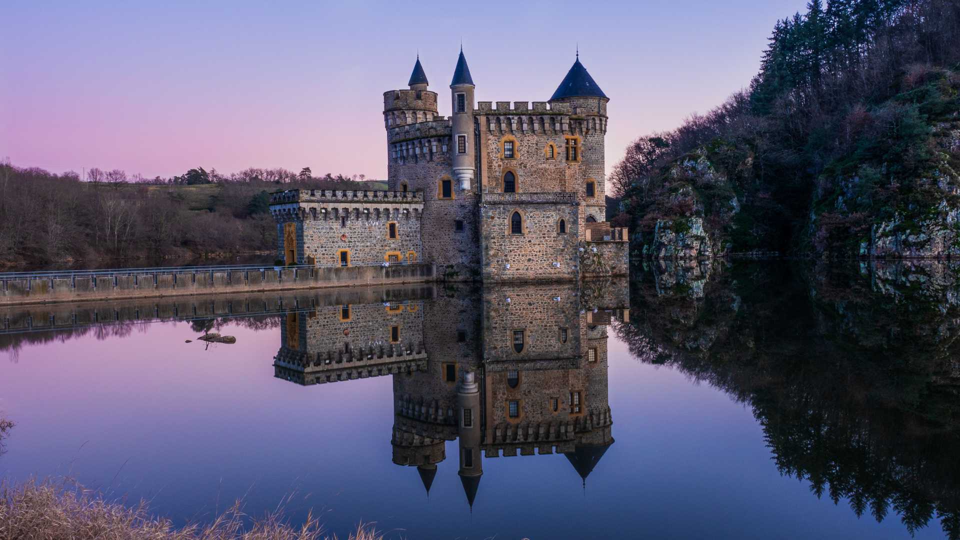 Chateau de la Roche in Saint-Priest-la-Roche, Frankrijk, weerspiegeld in kalm water bij zonsondergang met een paarse lucht.