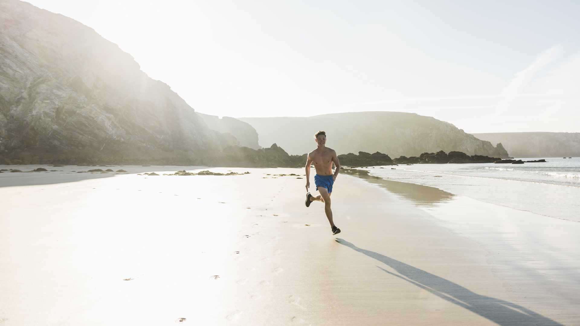 A young man runs shirtless on a serene beach of the Crozon Peninsula in France, bathed in soft sunlight.