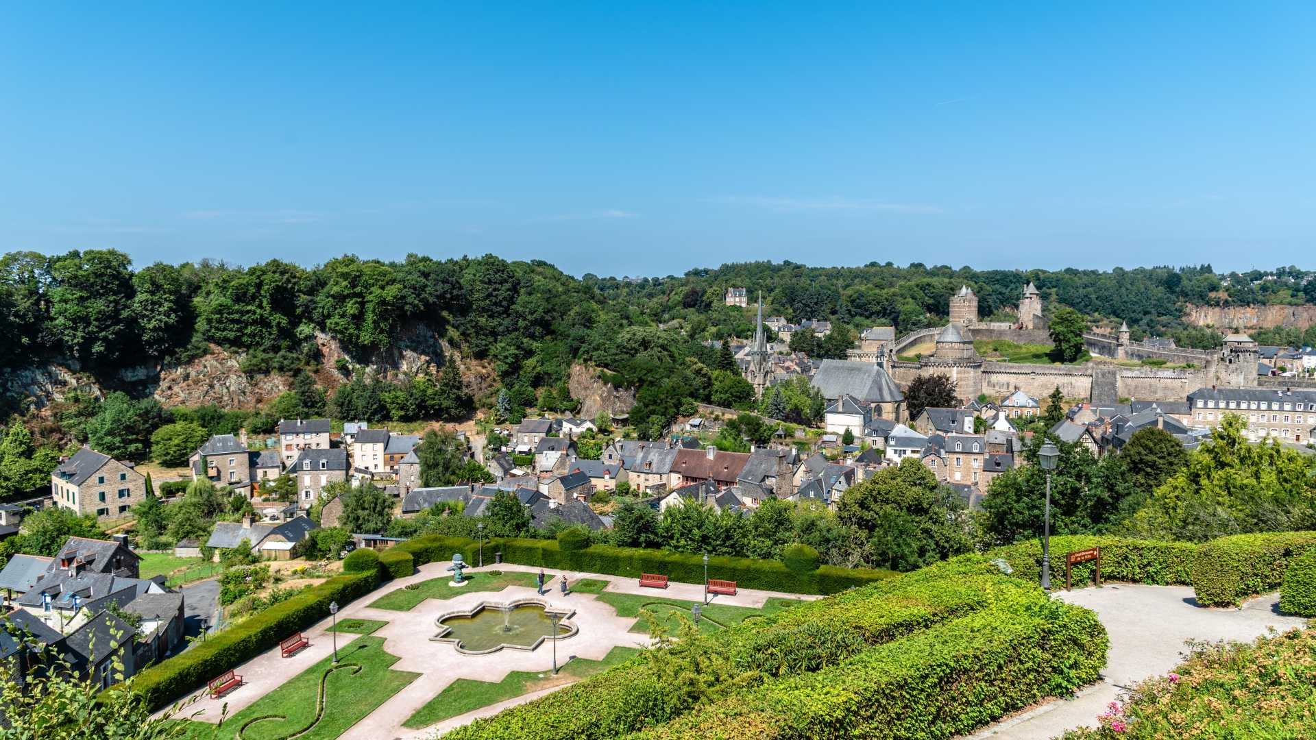 Medieval castle overlooks the scenic town of Fougères, with lush greenery under a clear summer sky.