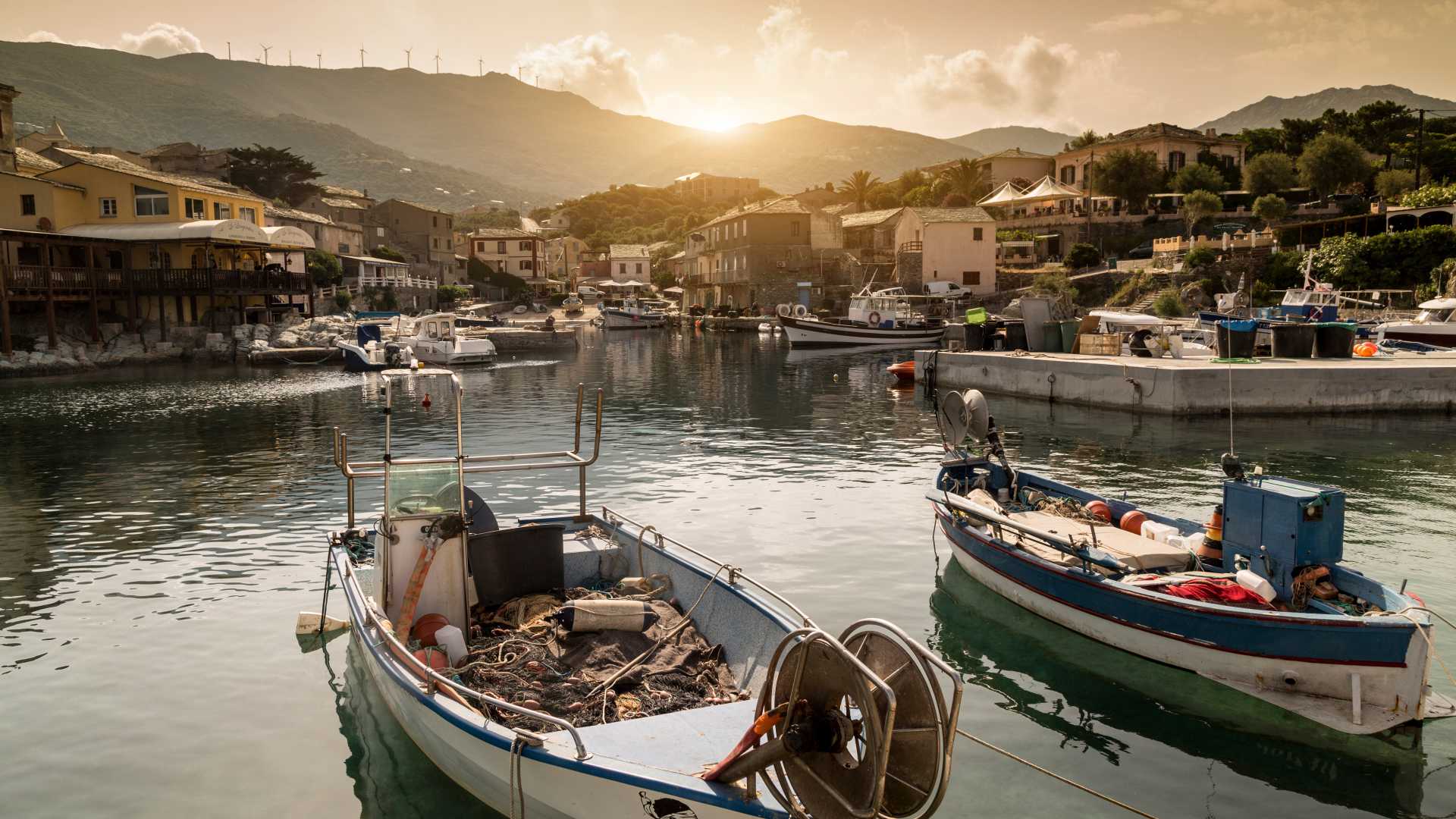 Fishing boats moored in the quiet harbour of Centuri, Corsica, with traditional houses in the background.