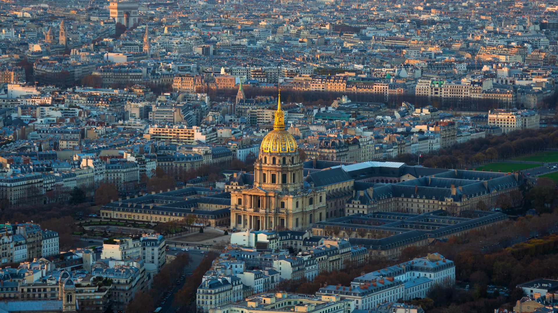 Aerial view of Les Invalides with its golden dome amidst the Parisian cityscape at dusk.