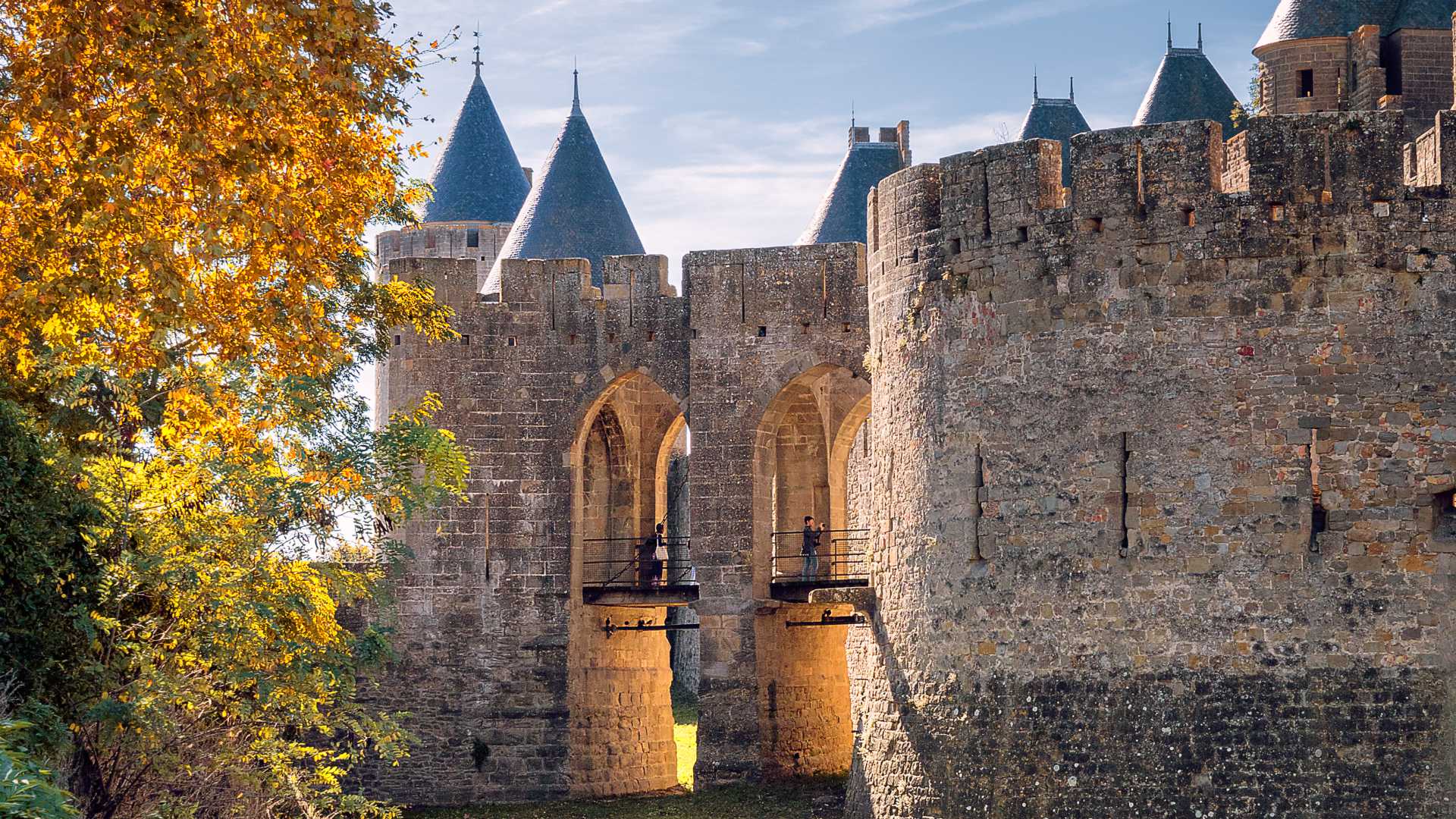 Entrée médiévale de la Porte Narbonnaise à Carcassonne, encadrée par des arbres d'automne et d'anciens murs de pierre.