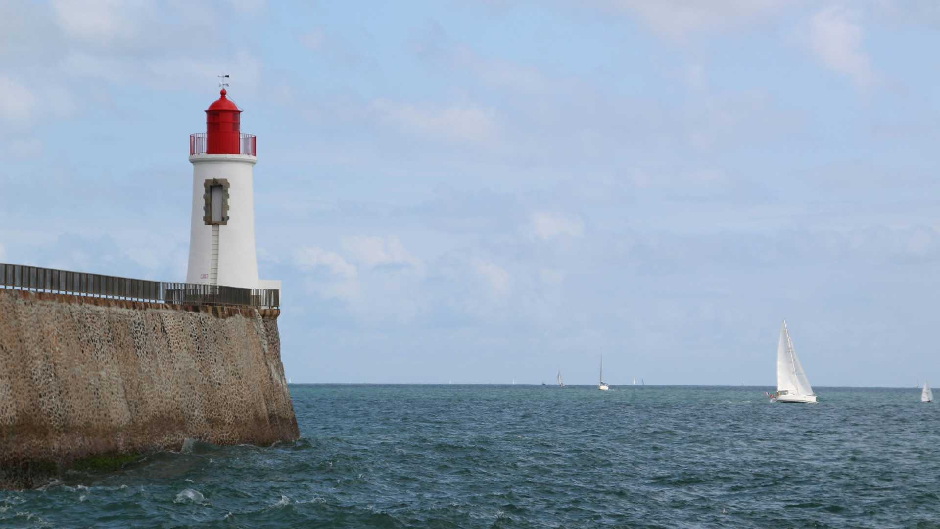 A white lighthouse with a red top at Les Sables-d'Olonne, France, overlooking a serene sea with sailboats.