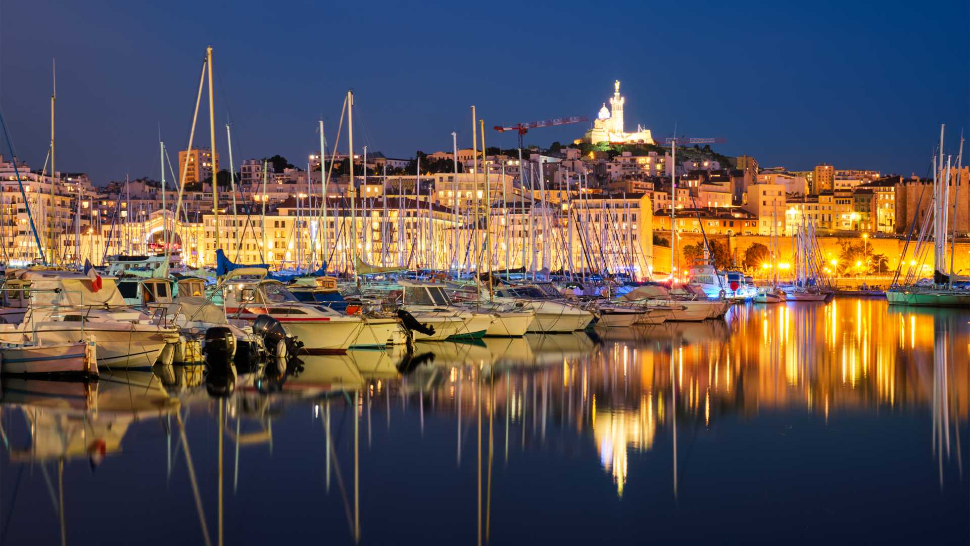 Les yachts se reflètent dans les eaux tranquilles du Vieux-Port de Marseille la nuit, avec la basilique Notre-Dame de la Garde illuminée en arrière-plan.
