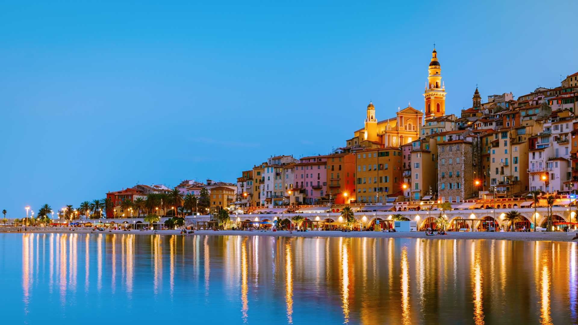 Colorful buildings of Menton reflect in tranquil waters at dusk, with a prominent church tower glowing warmly.