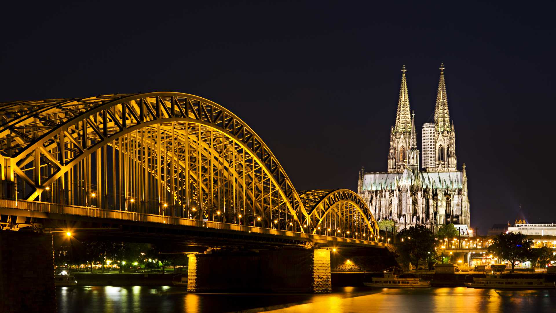 The illuminated Hohenzollern Bridge and Cologne Cathedral sparkle over the Rhine River at night.