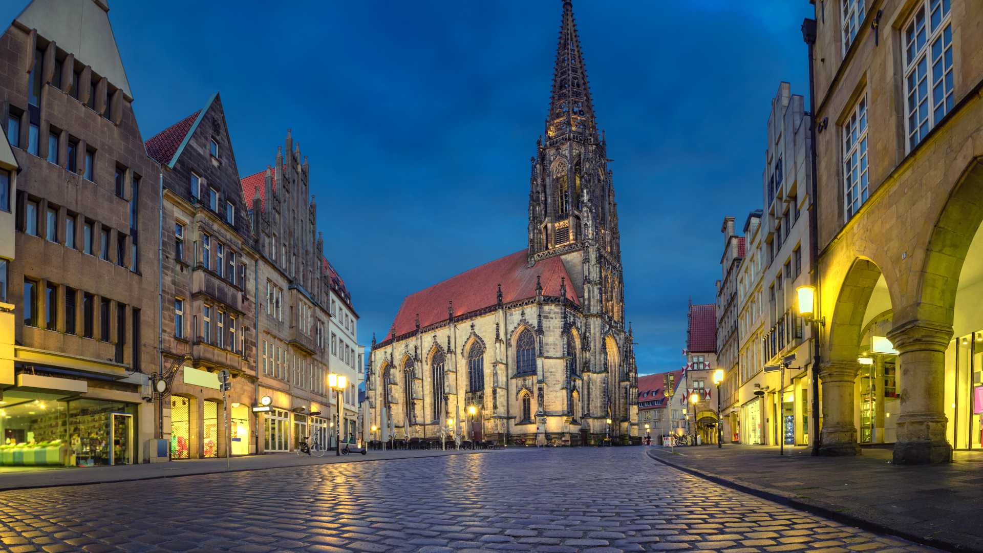 St. Lambert's Church in Münster illuminated at dusk, surrounded by historic buildings and cobblestone streets.