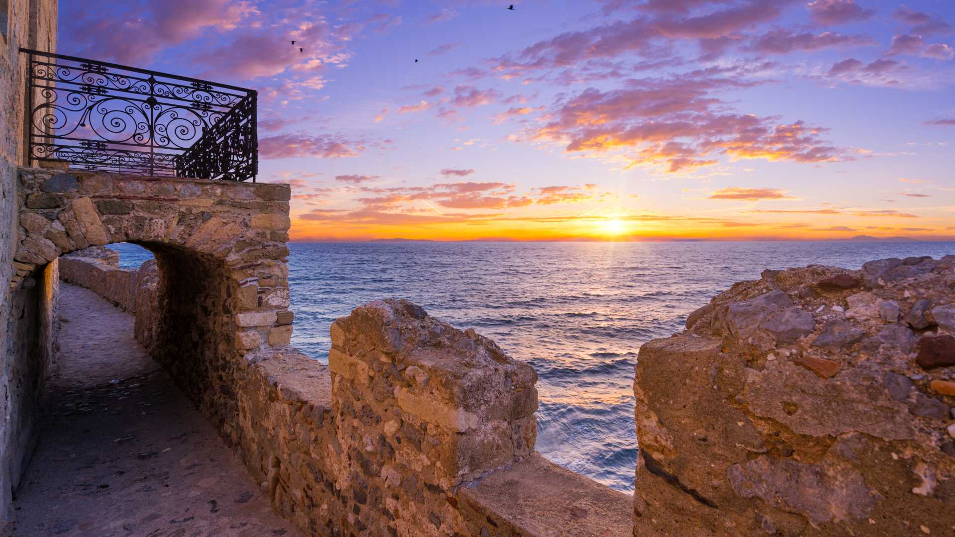 Zonsondergang vanaf de stenen muren van Monemvasia, met een historische boog en balkon met uitzicht op de Egeïsche Zee.