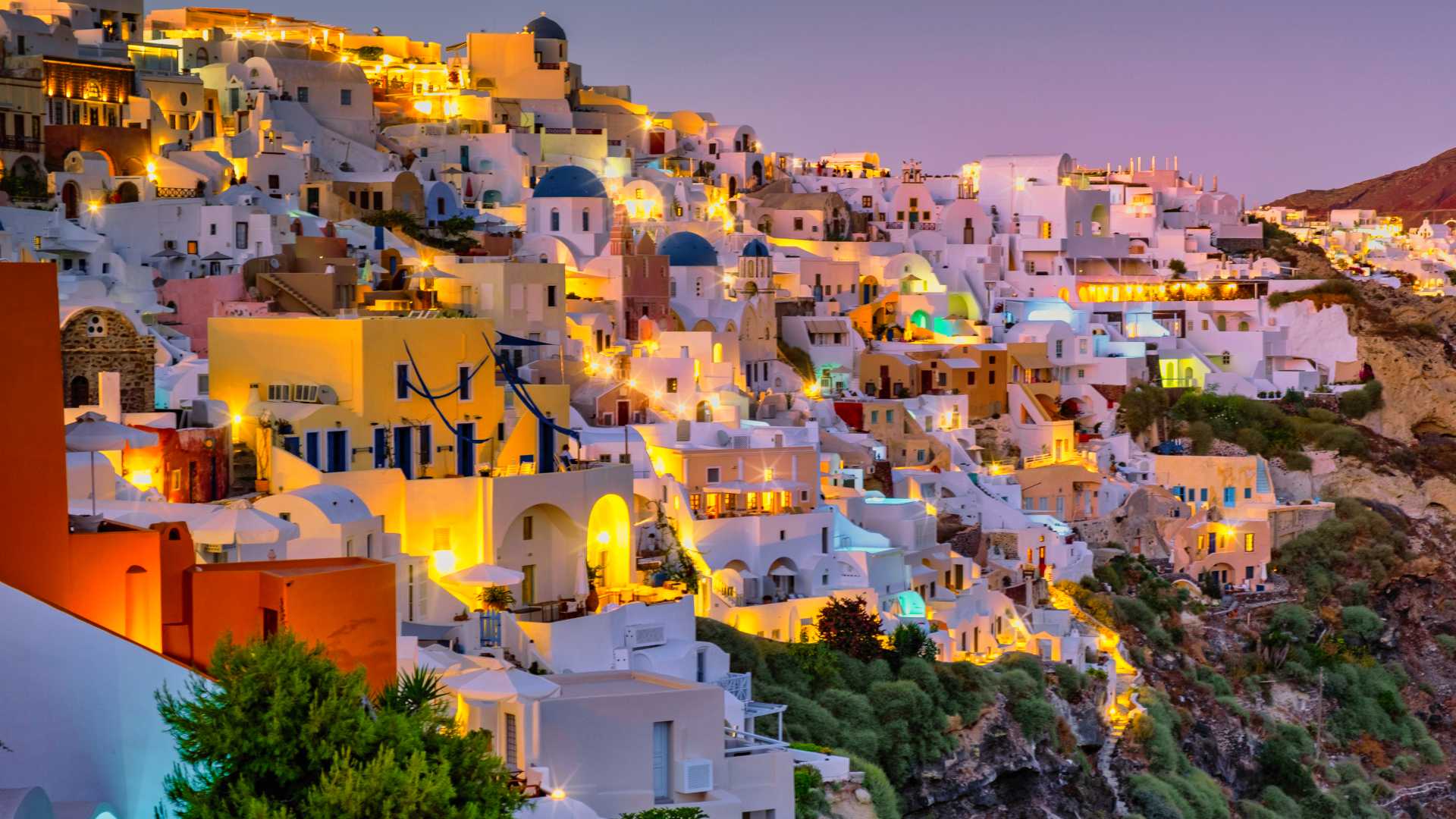 Whitewashed buildings of Oia, Santorini, glow warmly at sunset, with domed churches and a serene evening sky.