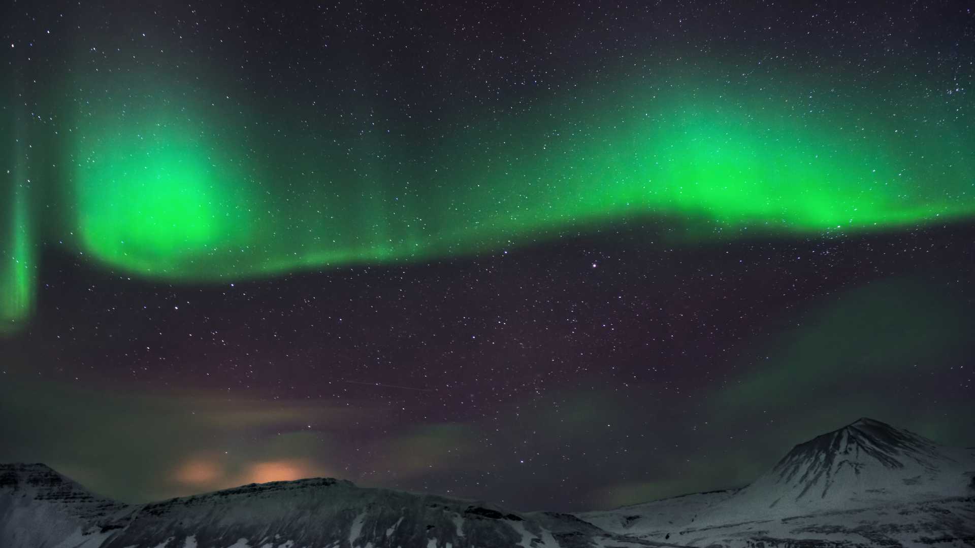 Green aurora borealis dances above snow-capped mountains under a starry night sky in Iceland.