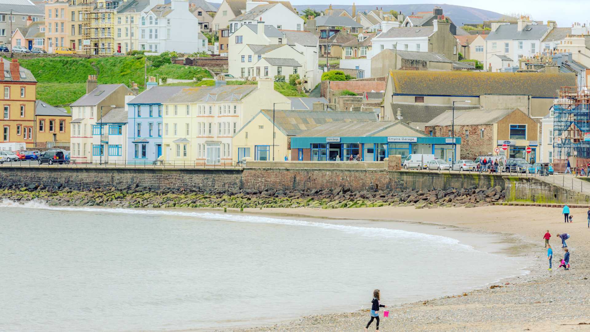 Kleurrijke gebouwen langs de kustlijn van een zandstrand in Peel, Isle of Man, met mensen die een wandeling maken.