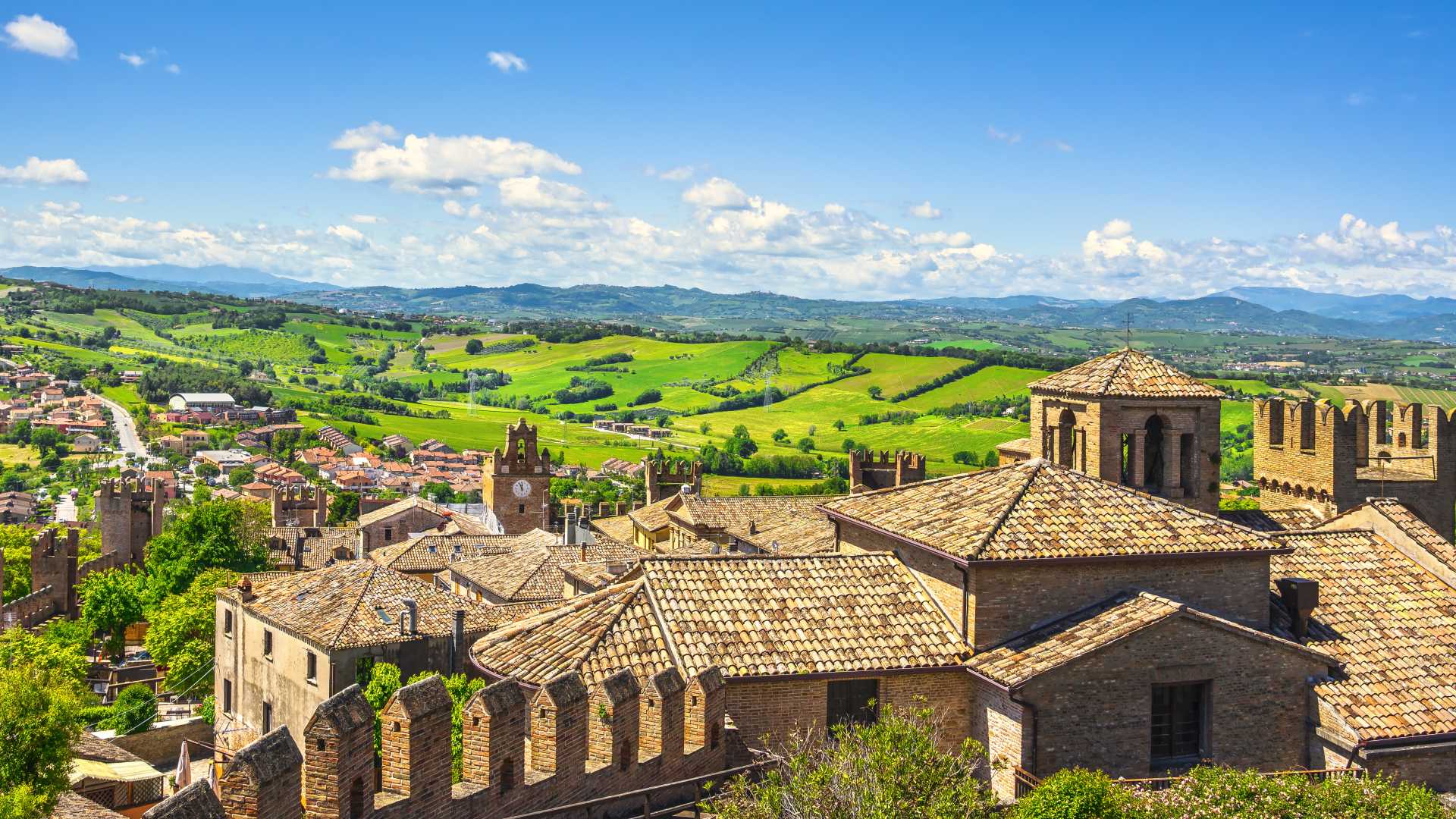 A panoramic view of Gradara's medieval rooftops and the surrounding green hills under a bright, clear sky in Marche, Italy.