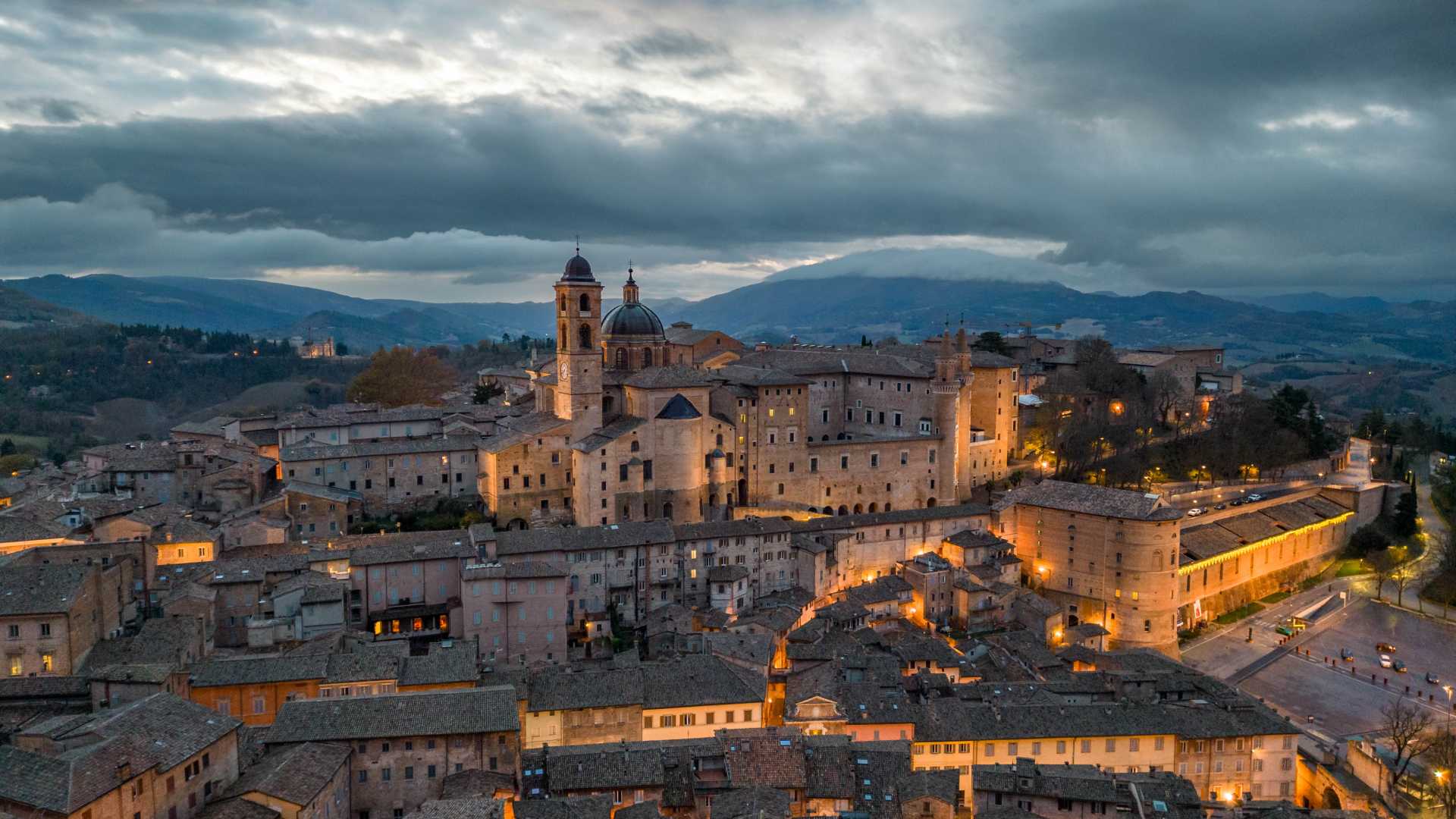 Urbino's historic buildings glow softly as the sun rises, set against a backdrop of rolling hills.