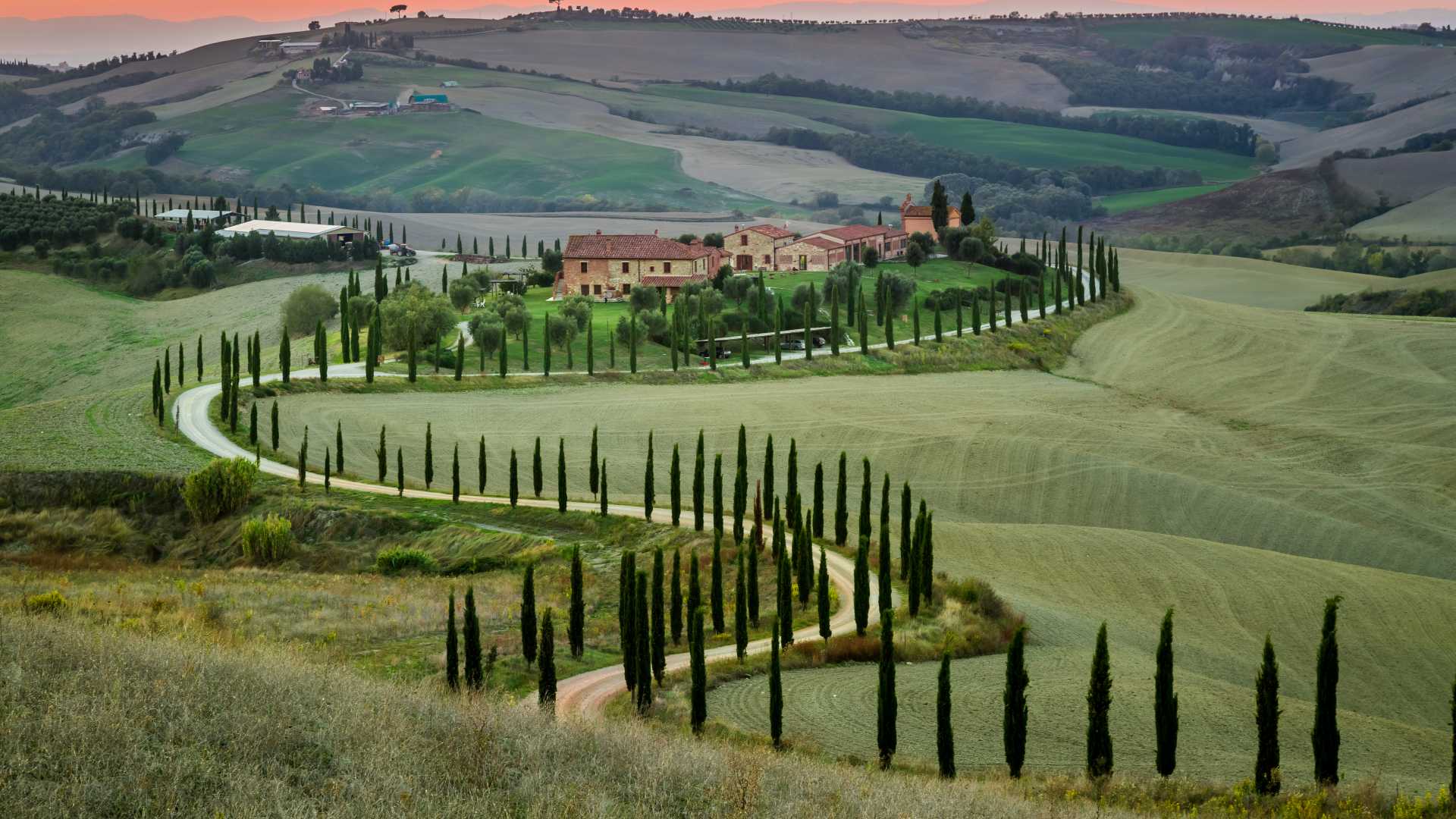 Kronkelende weg omzoomd met cipressen leidt naar een Toscaanse villa, genesteld in glooiende groene heuvels, gekust door de ondergaande zon.