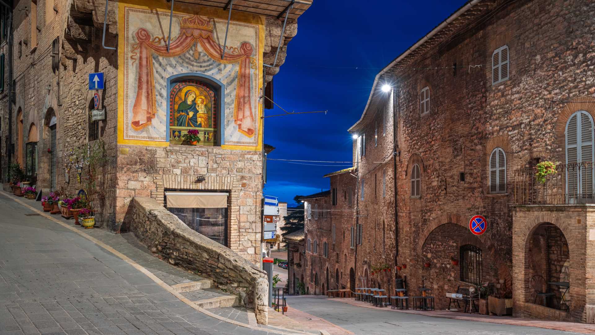 Historische middeleeuwse straten van Assisi, Italië, in de schemering, met stenen gebouwen en een kleurrijke religieuze muurschildering.