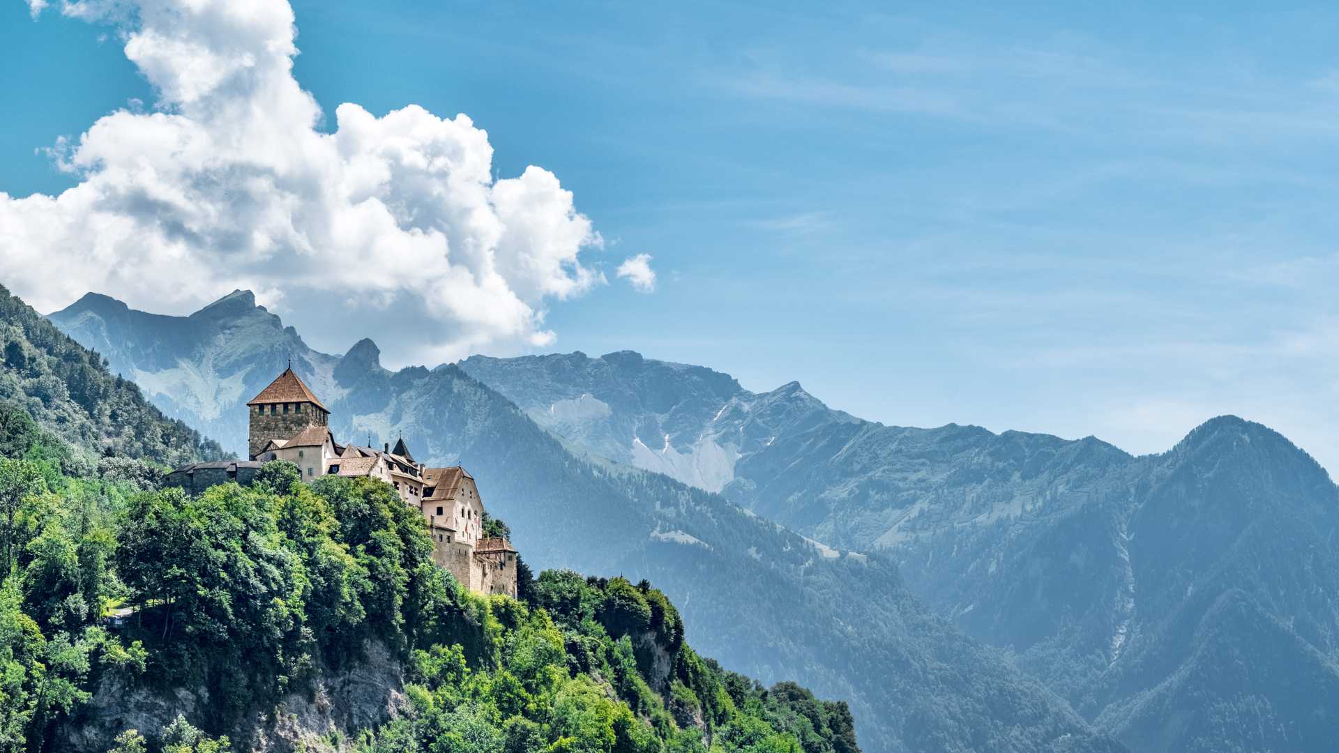 Il Castello di Vaduz arroccato su una lussureggiante collina con le Alpi sullo sfondo sotto un cielo azzurro brillante nel Liechtenstein.