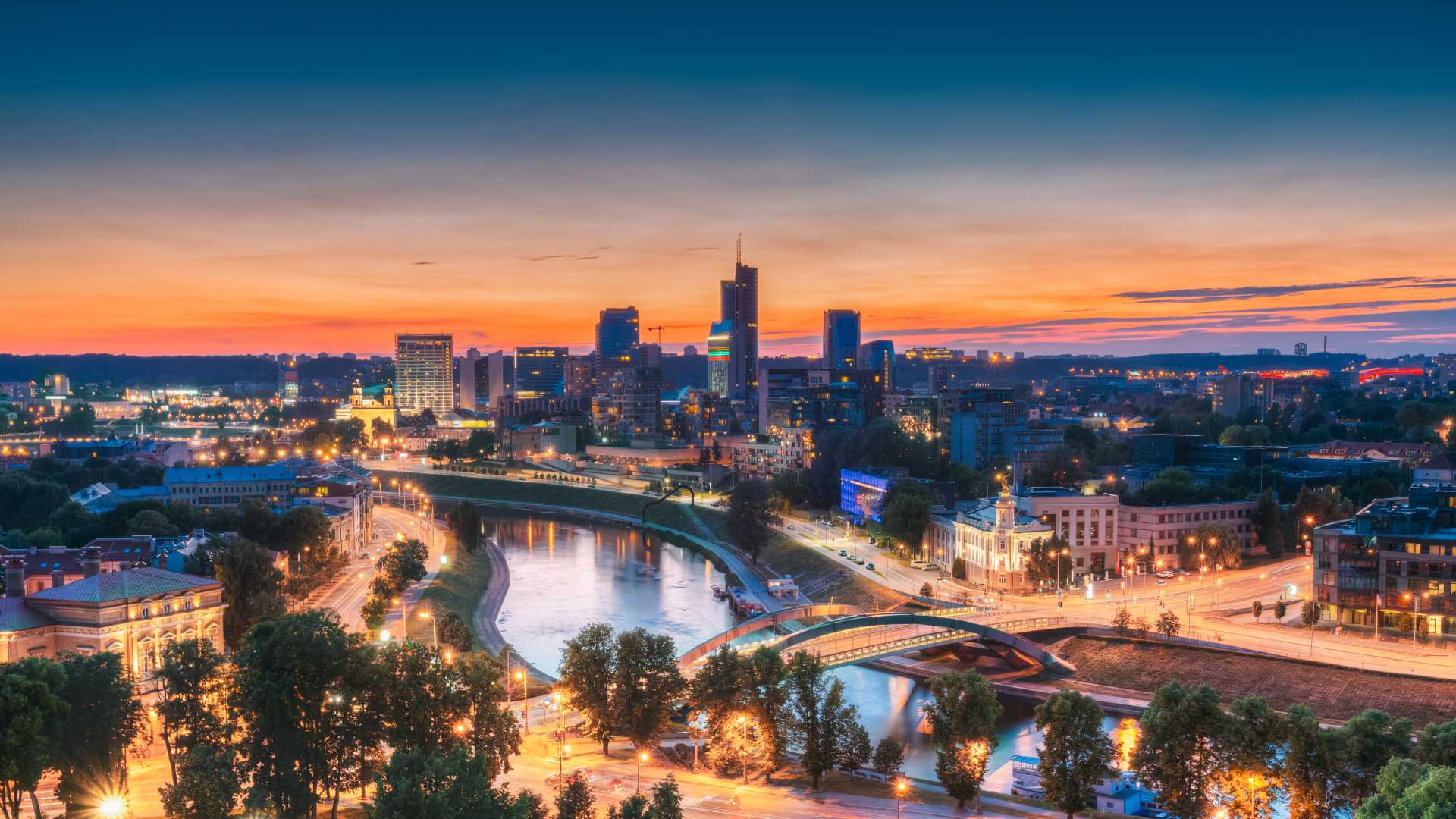 Vilnius cityscape at sunset, featuring modern skyscrapers and the Neris River illuminated at night.