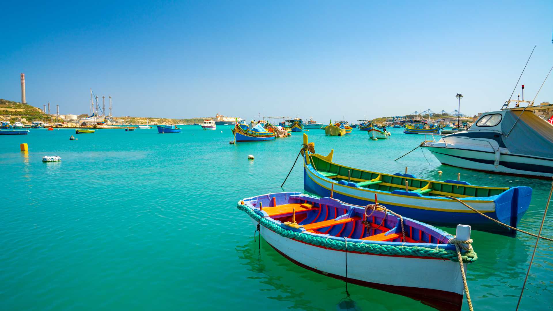Colorful traditional luzzu boats float in Marsaxlokk Harbor, Malta, under a clear blue sky.