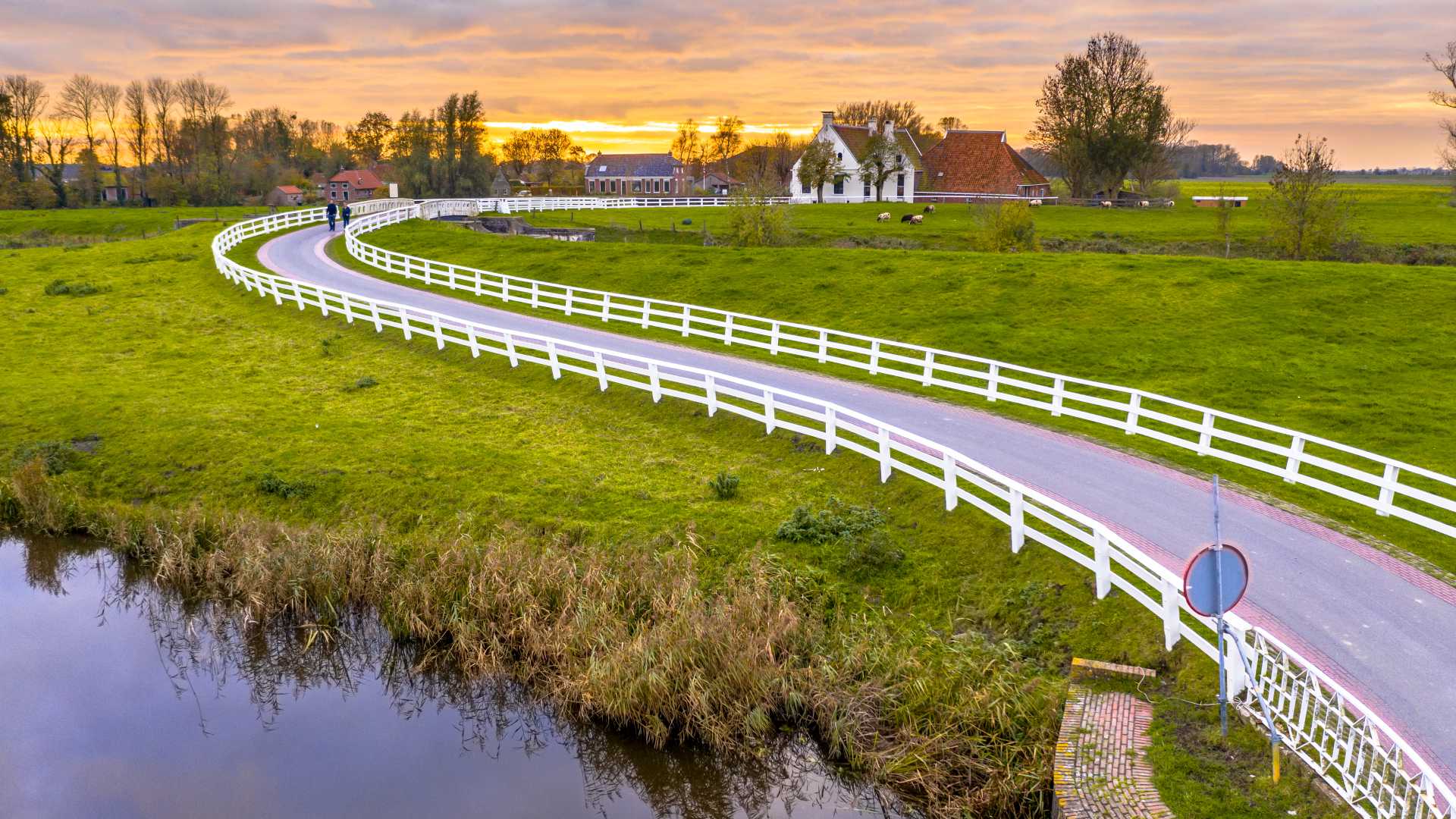 A winding road with white fences through green fields and historic houses at sunset in Groningen.