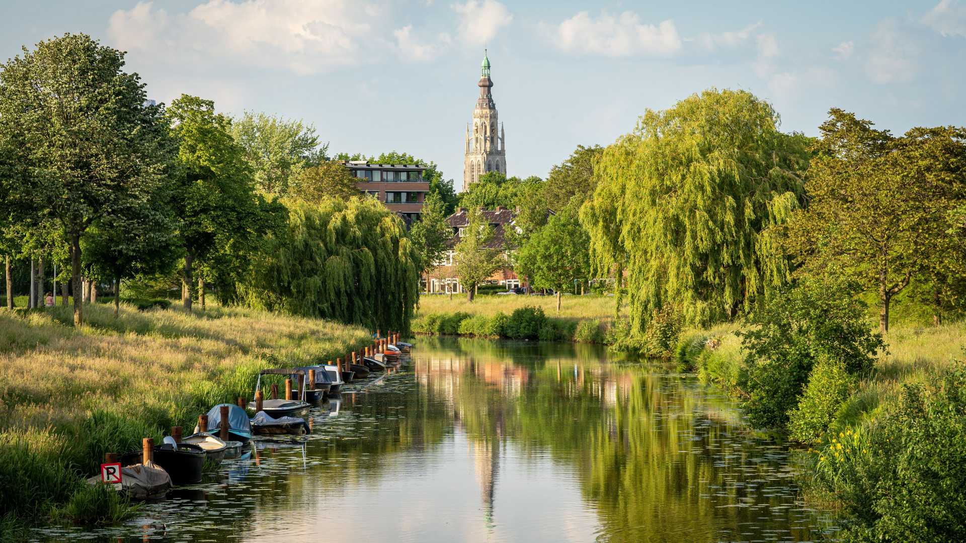 De iconische Grote Kerk van Breda torent boven groene landschappen en een rustig kanaal met kleine boten uit.