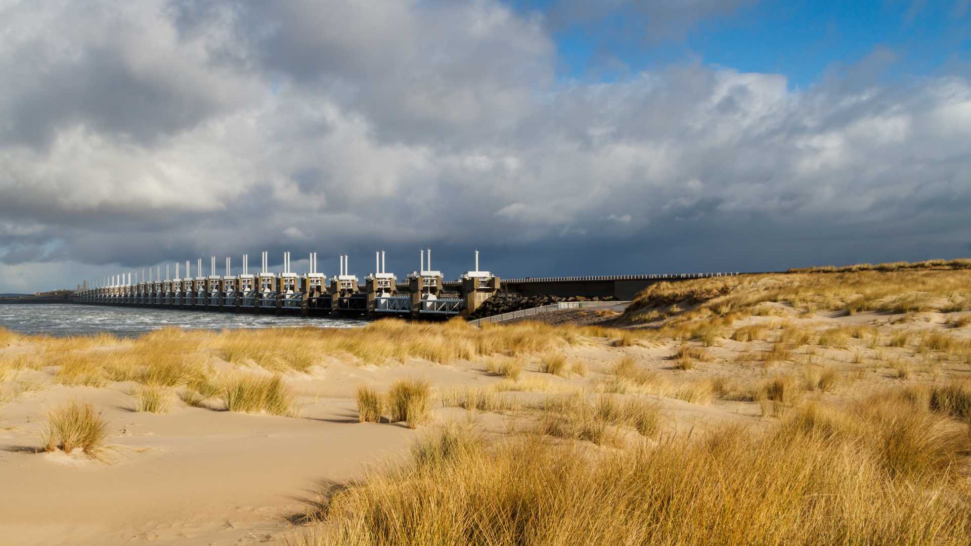View of the Eastern Scheldt Storm Surge Barrier under a cloudy sky, framed by golden sand dunes and beach grass.