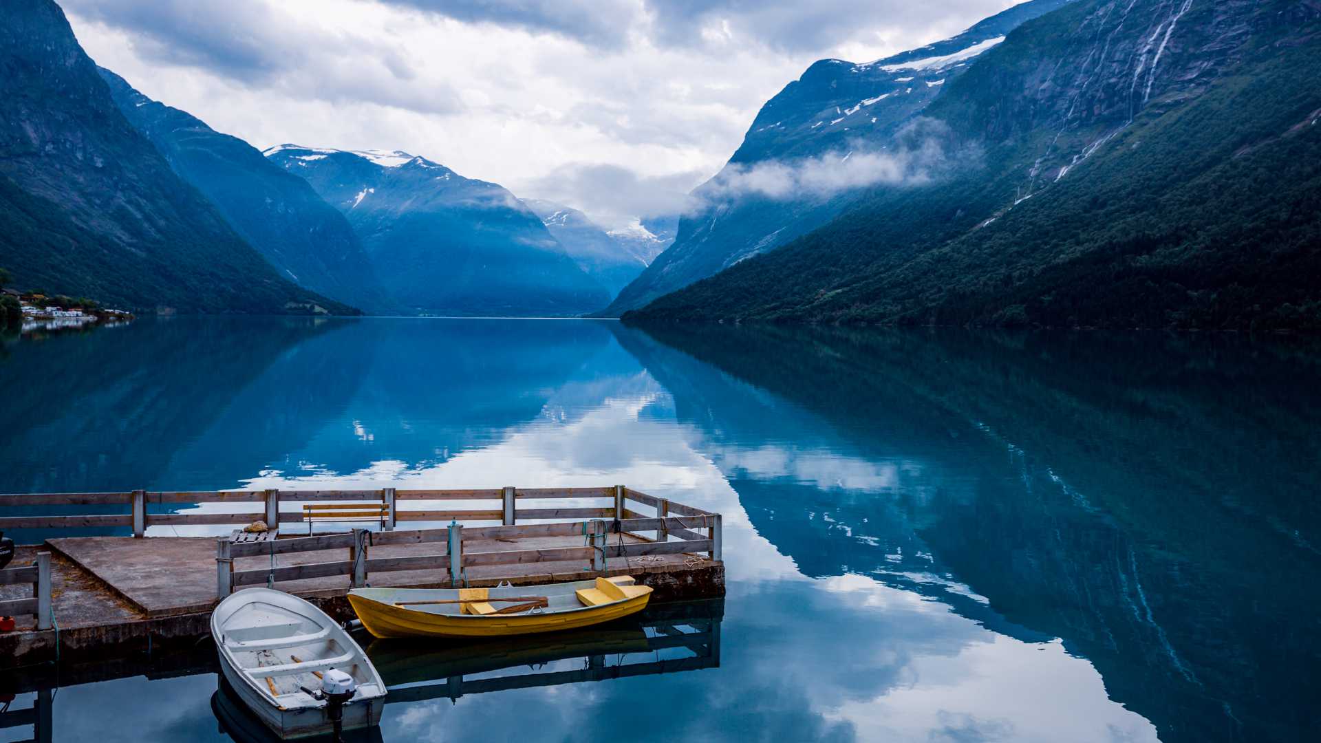 Tranquilo lago Lovatnet en Noruega con barcos atracados, rodeado de imponentes montañas y reflejos tranquilos.