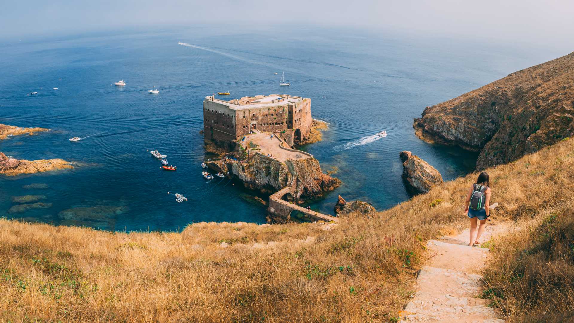 Un escursionista scende un sentiero erboso dorato verso lo storico Forte di São João Baptista, una fortezza in pietra costruita su una piccola isola nelle limpide acque blu dell'Arcipelago delle Berlengas. Barche punteggiano l'oceano calmo intorno al forte e alle scogliere rocciose.