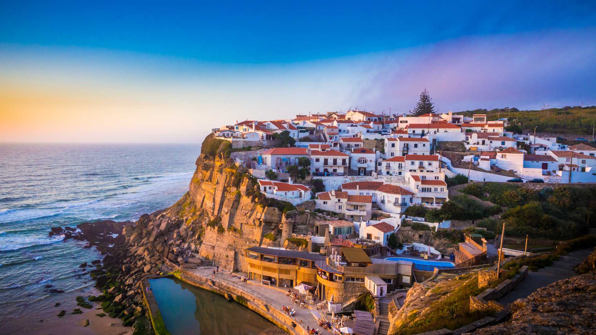 Whitewashed houses with red roofs perched on a cliff, overlooking the ocean in Azenhas do Mar, Portugal.