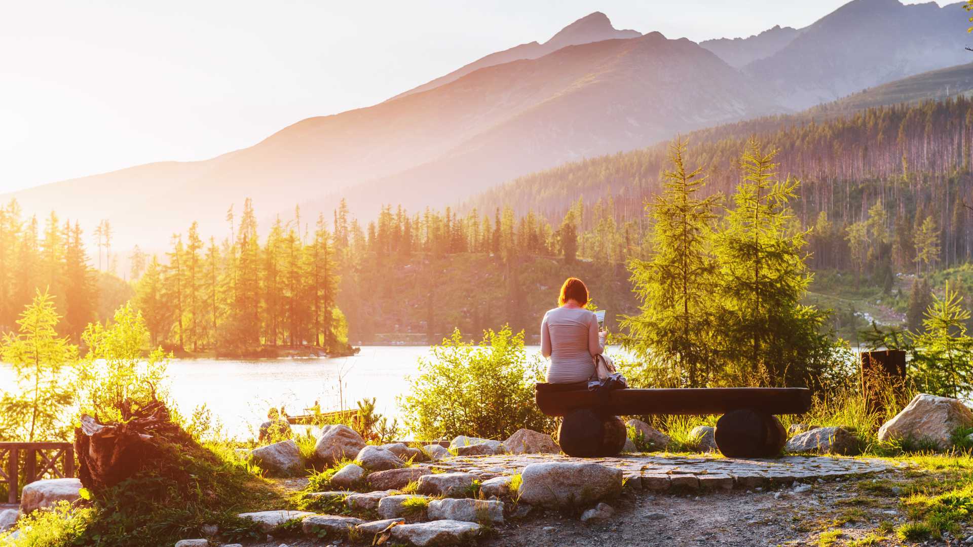A serene view of Lake Strbske Pleso with a person sitting on a bench, surrounded by greenery and mountains.