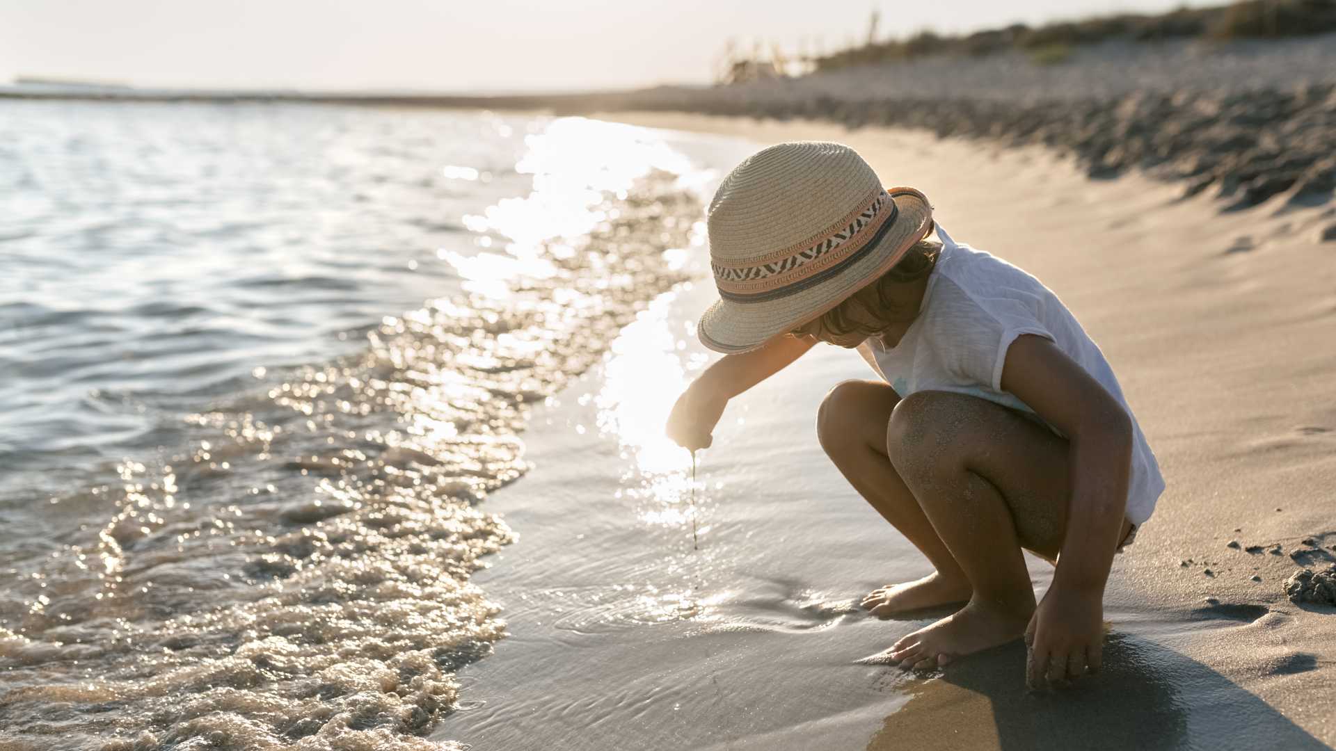Een klein meisje met een zonnehoed speelt bij zonsondergang aan de glinsterende oever van een strand in Menorca, Spanje.