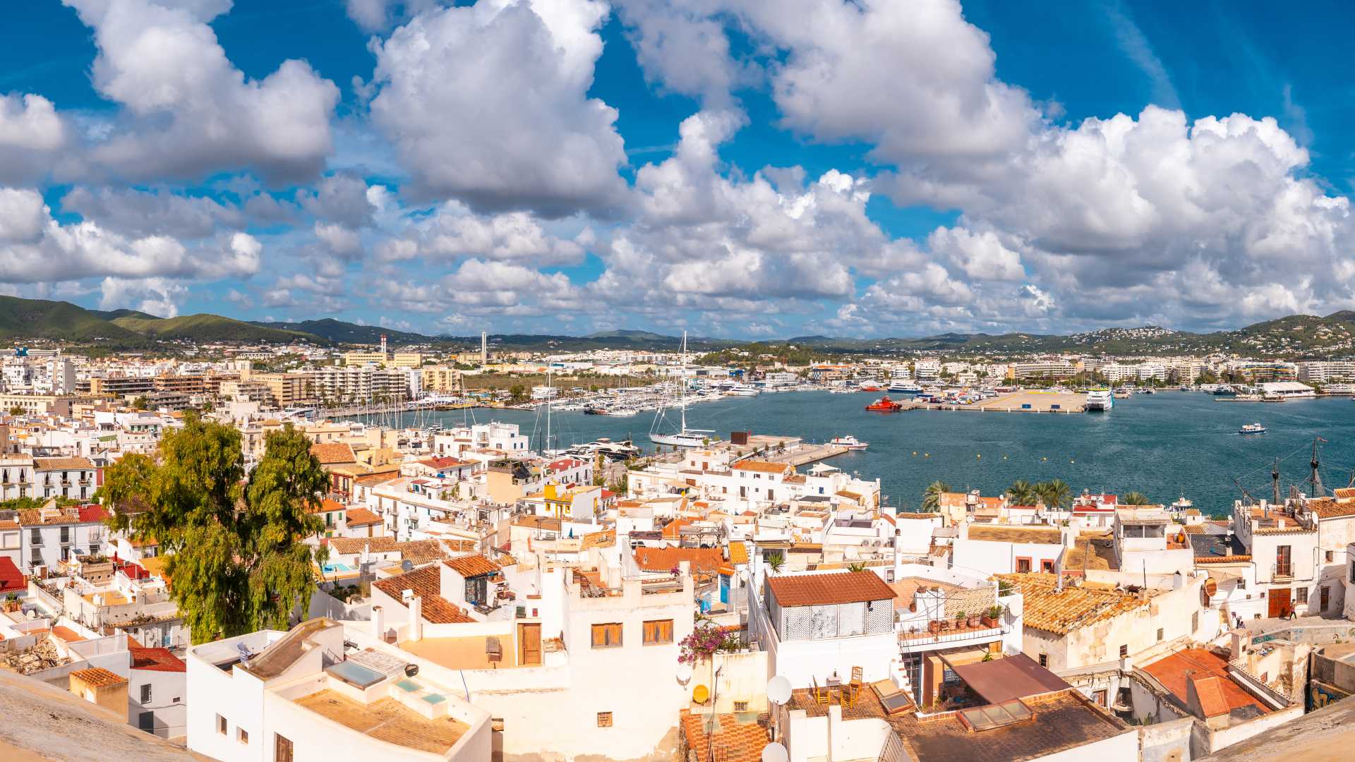 Een weids uitzicht op de historische oude stad en de levendige haven van Ibiza, omlijst door de kasteelmuren onder een blauwe, met wolken bezaaide hemel.