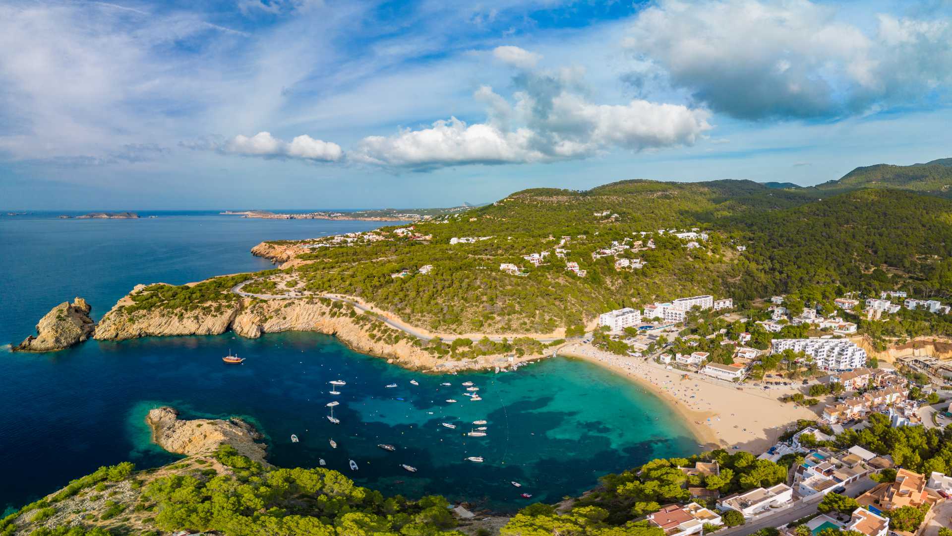 Aerial view of Cala Vadella's turquoise waters, sandy beach, and green hills in Ibiza, Spain.