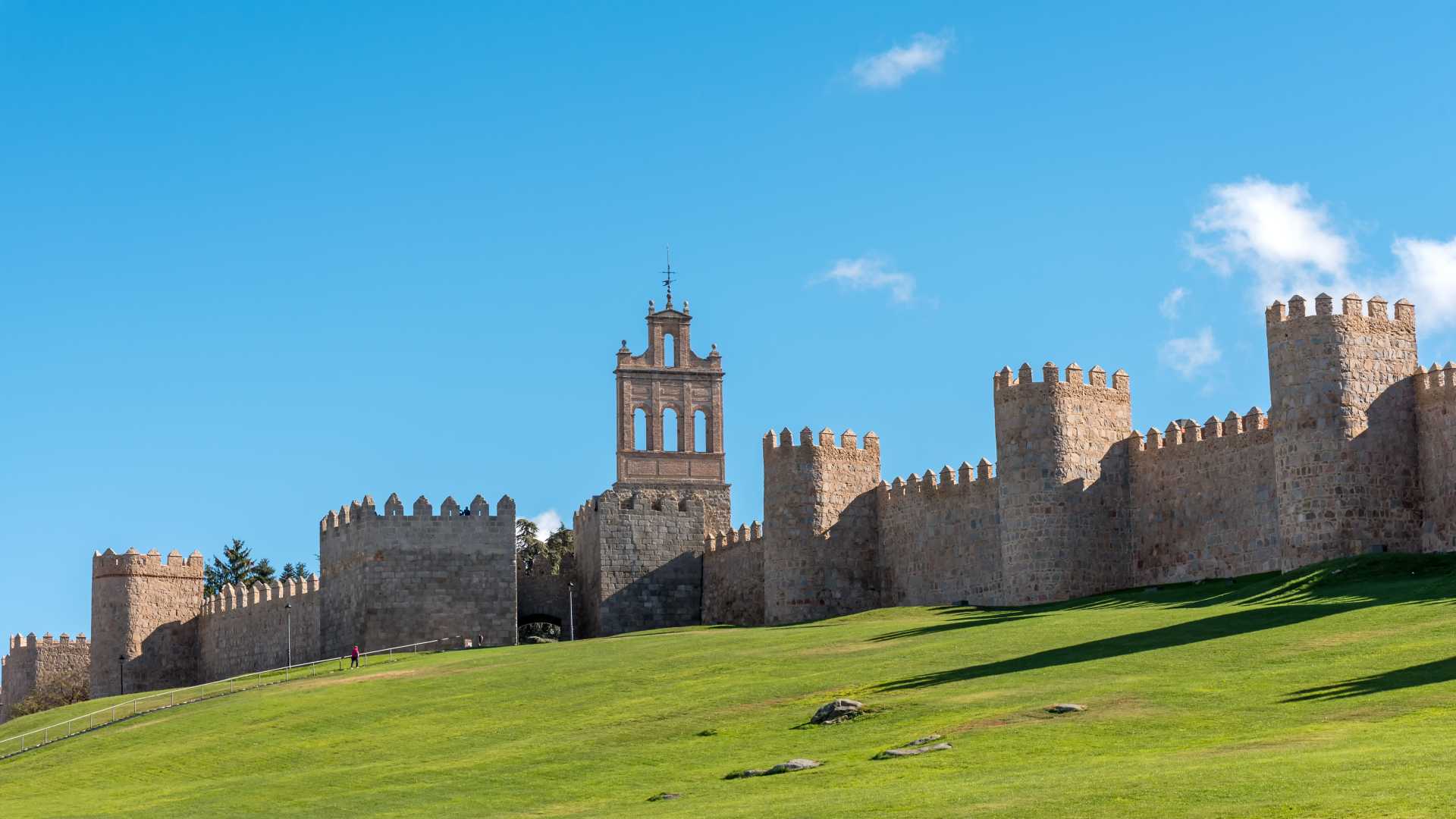 Middeleeuwse stadsmuur van Ávila met kantelen en een toren, tegen een heldere blauwe lucht en groen gras.
