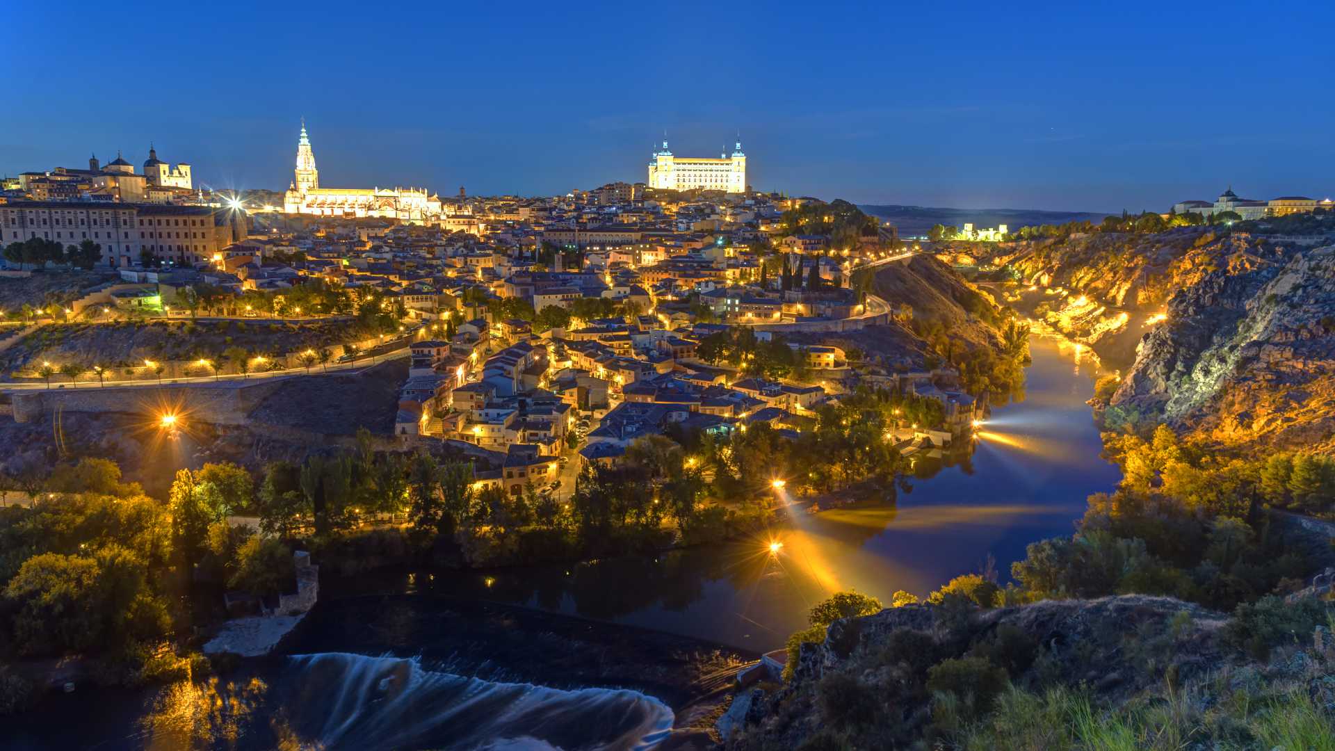 Toledo's historic city at night, featuring the Alcázar, Cathedral, and the Tagus River under a twilight sky.