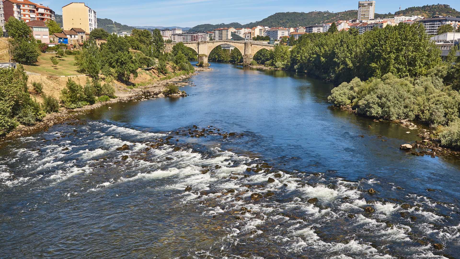 De oude Romeinse brug overspant sierlijk de rivier de Miño in Ourense, Spanje, omlijst door weelderig groen en de skyline van de stad.