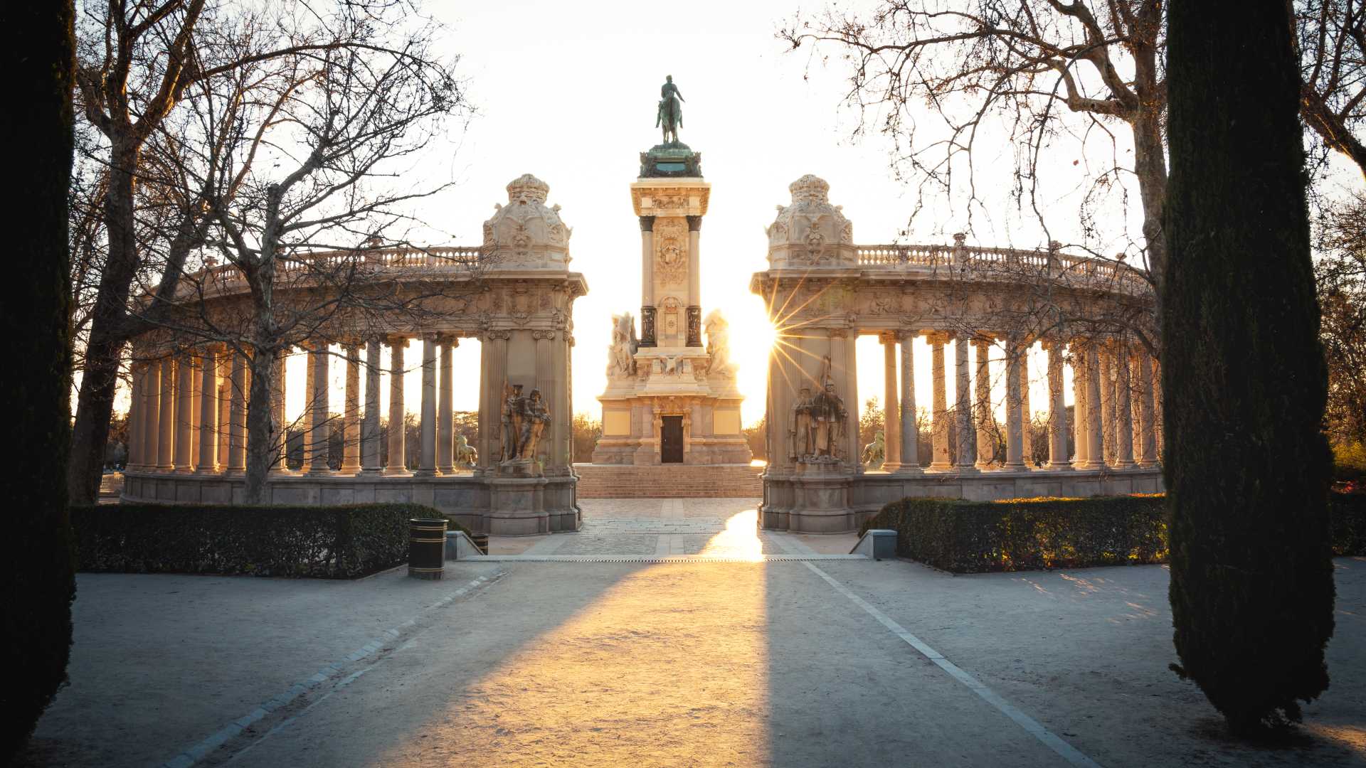 Zonovergoten monument voor Koning Alfonso XII in het El Retiro Park, Madrid, omlijst door kale bomen en grote zuilen.