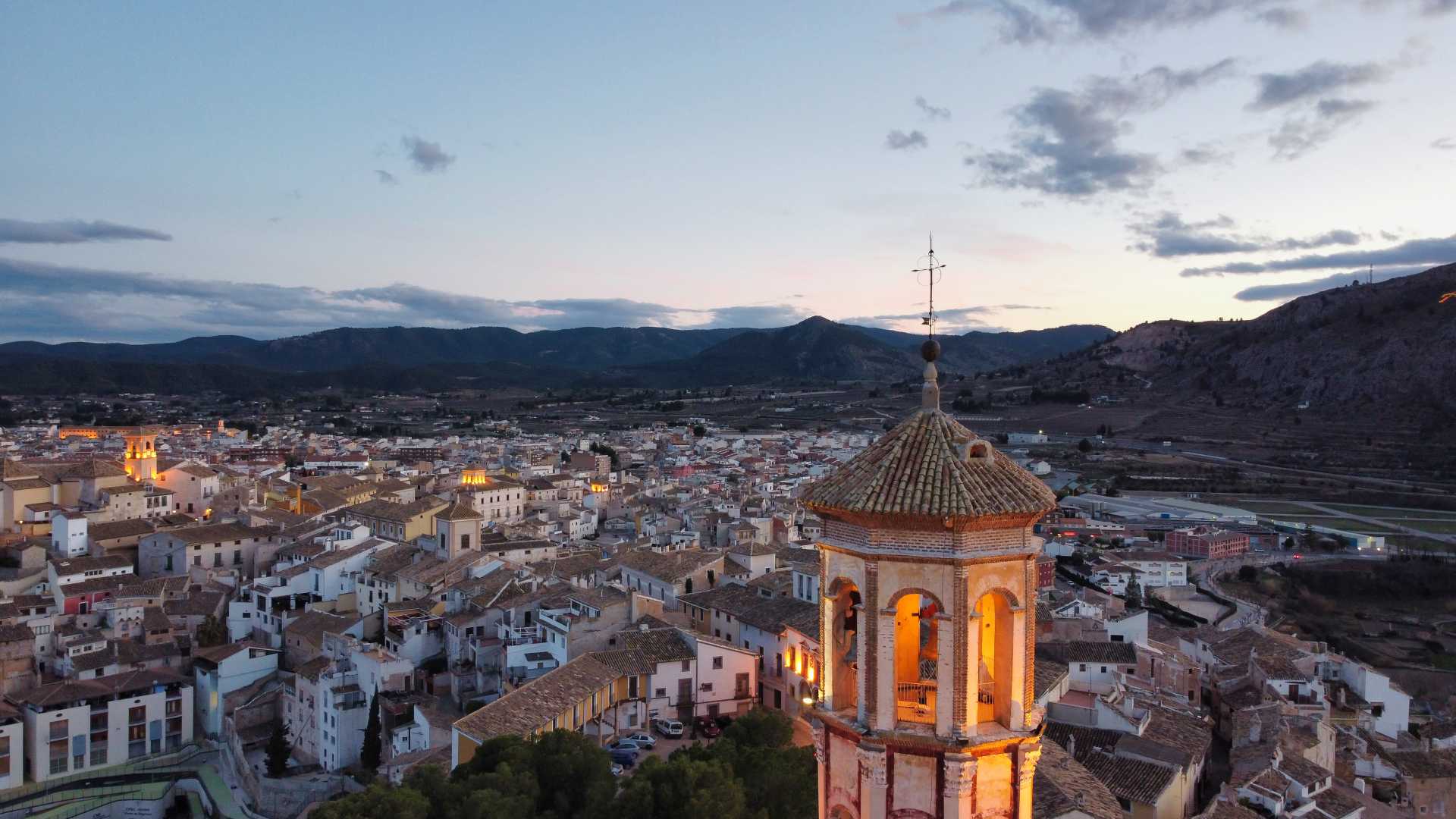 Avondzicht op Cehegín, Murcia, met een verlichte kerktoren en daken tegen een bergachtige achtergrond.