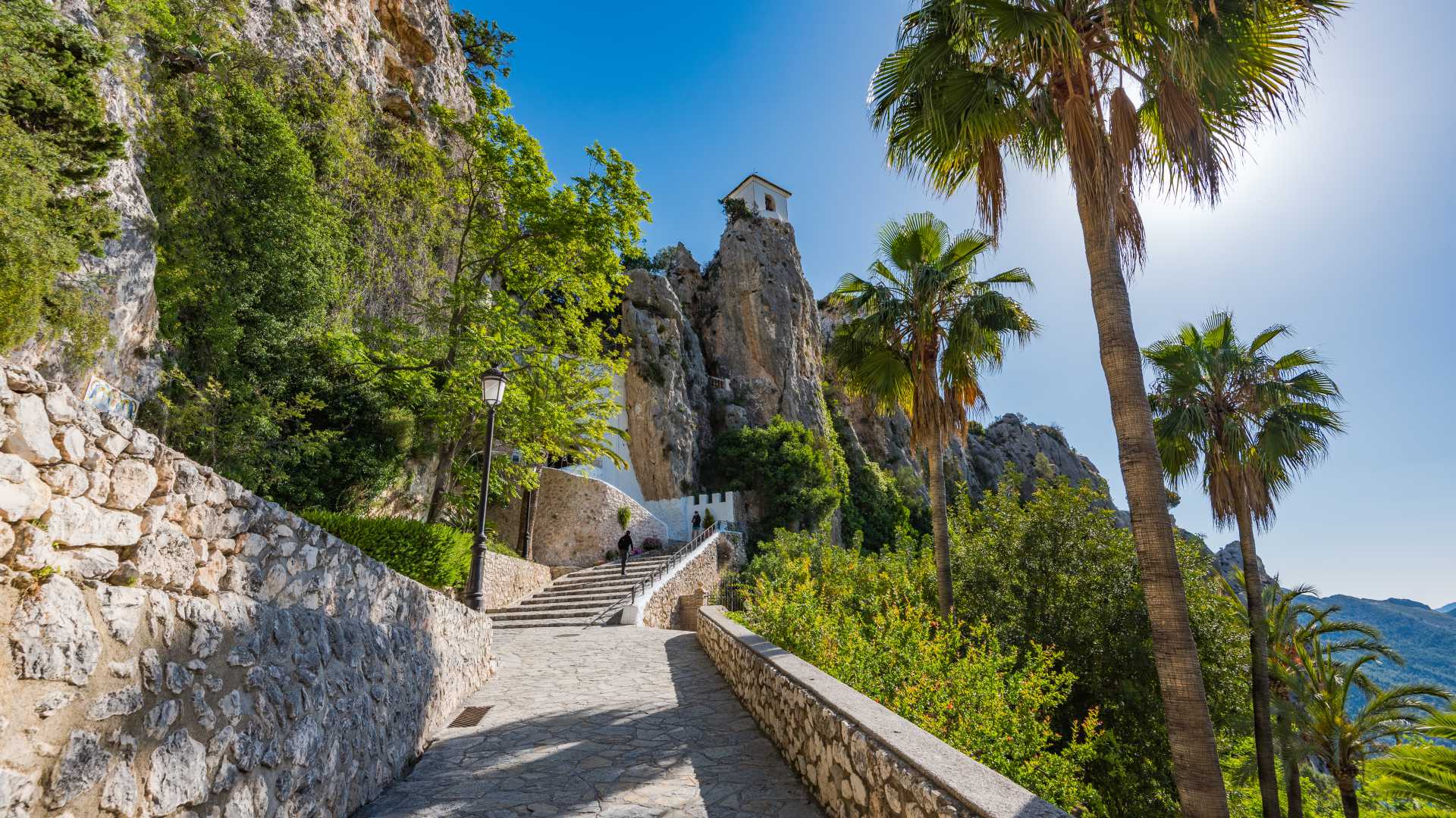 Een stenen pad leidt naar het kasteel van Guadalest, gelegen op een rotsachtige klif, omgeven door groen en palmbomen in Alicante, Spanje.