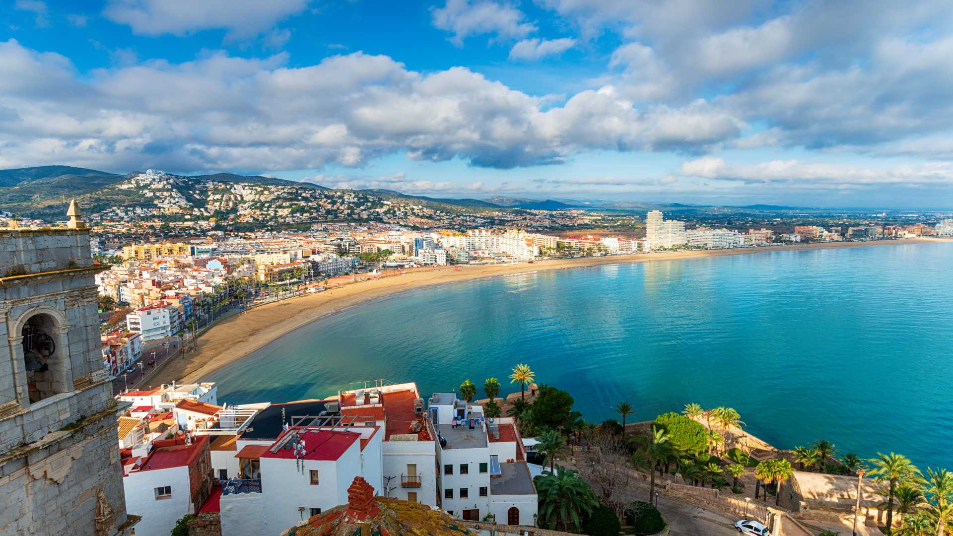 Panoramisch uitzicht op de daken van Peñíscola, het gouden strand en het turquoise water van de Middellandse Zee met bergen op de achtergrond.